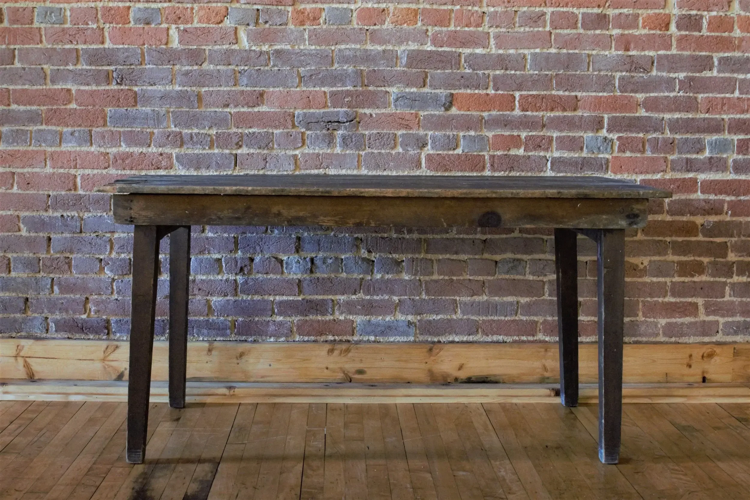 A rustic wooden table with black metal legs placed against a red brick wall in a room with wooden flooring.