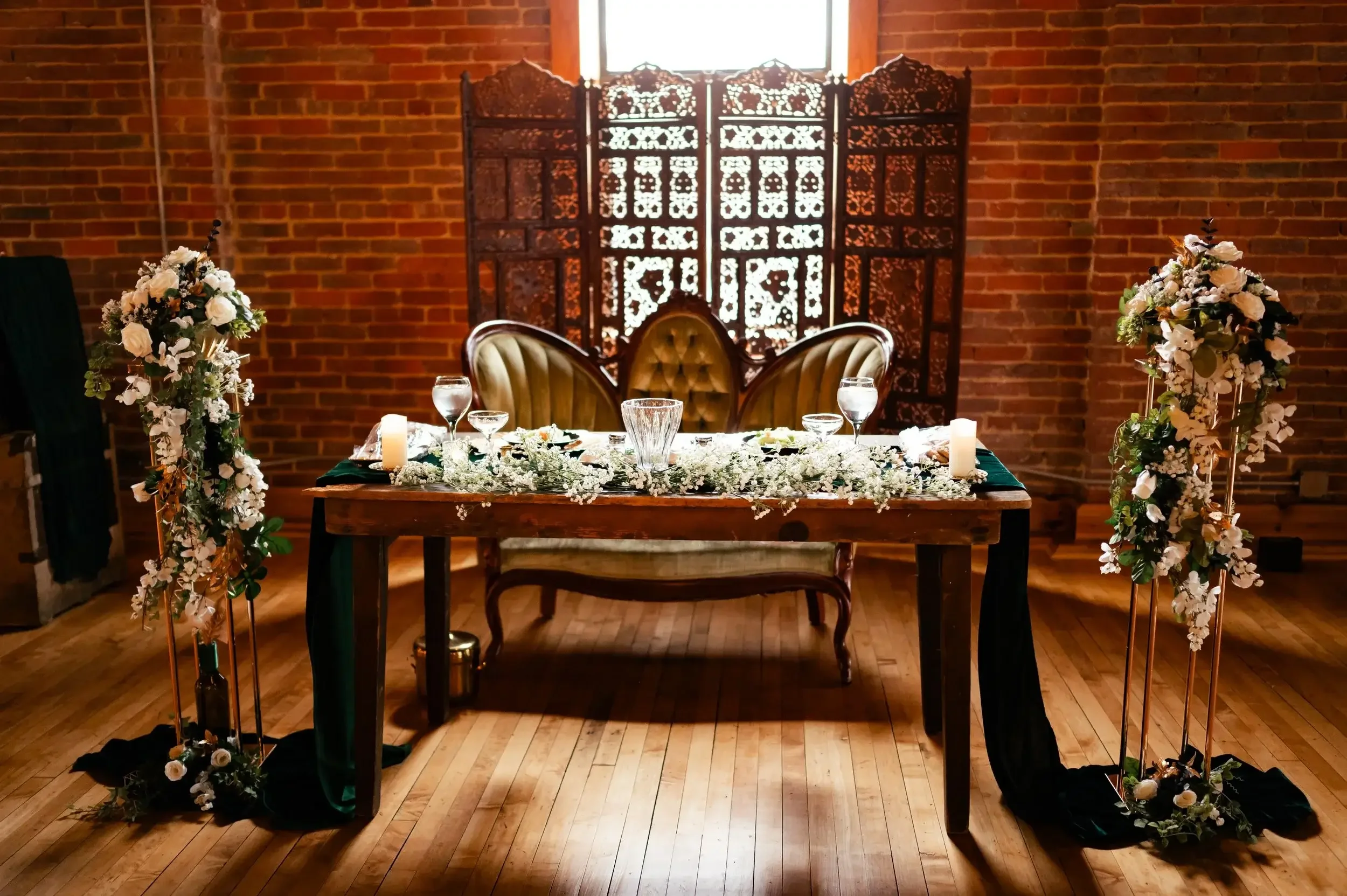 Elegant wedding reception table with floral arrangements, candles, and glassware in front of a vintage folding screen with a brick wall background.