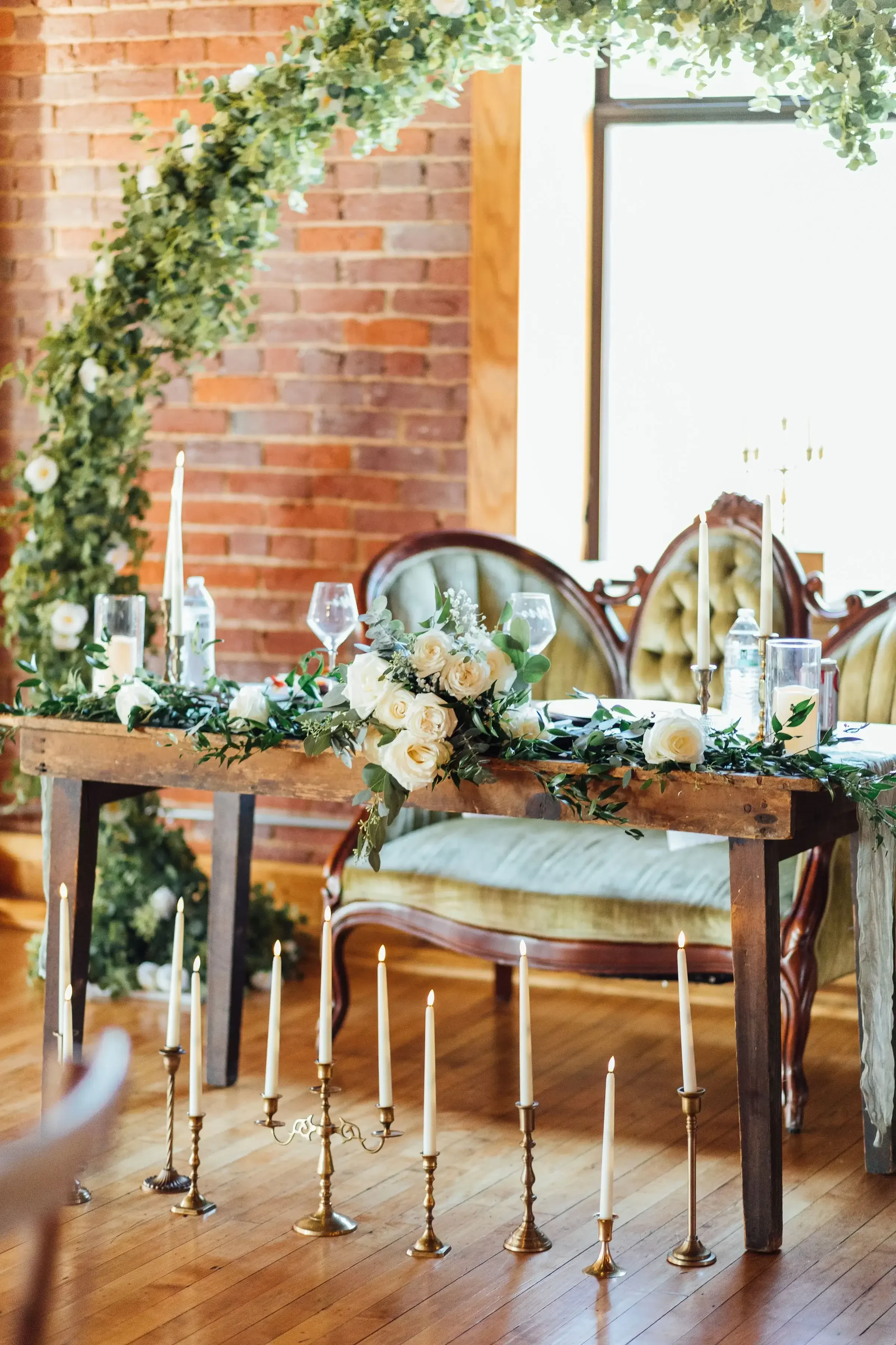 A decorated table with floral arrangements, candles, and glassware, set for a wedding or special event, with vintage chairs and a brick wall in the background.