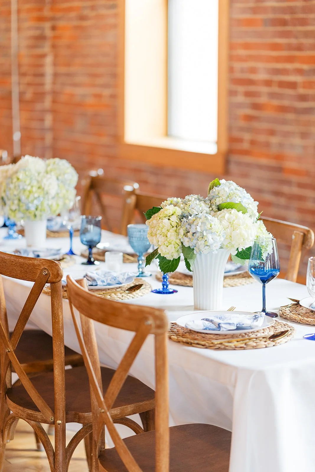 A table set with white flowers in vases, blue glasses, woven placemats, and napkins with blue patterns in a brick-walled room.