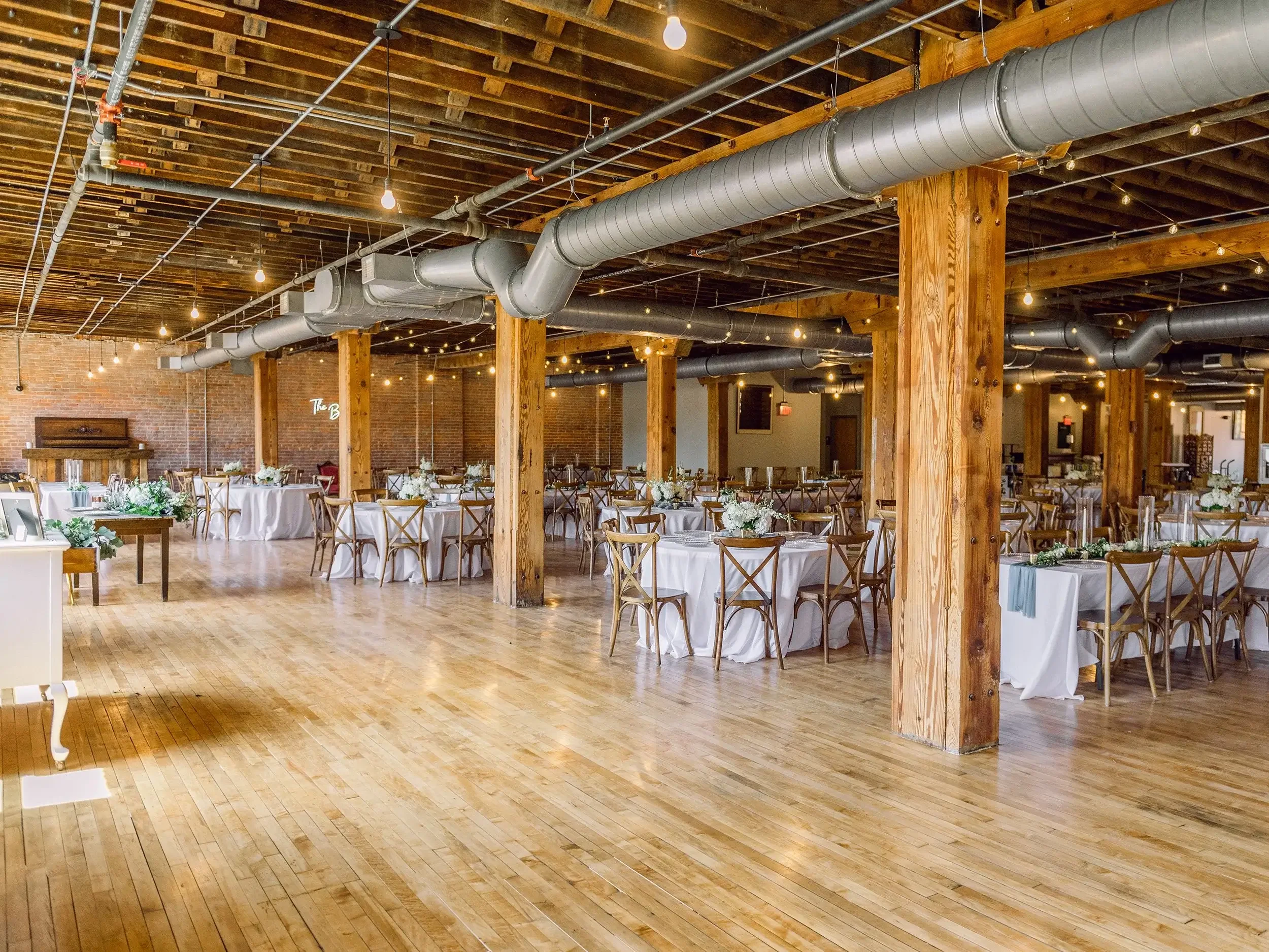 A spacious event hall with wooden flooring, wooden support beams, and exposed brick walls. The ceiling has industrial-style ductwork and hanging string lights. Several round tables are set with white tablecloths and floral centerpieces, and wooden ch