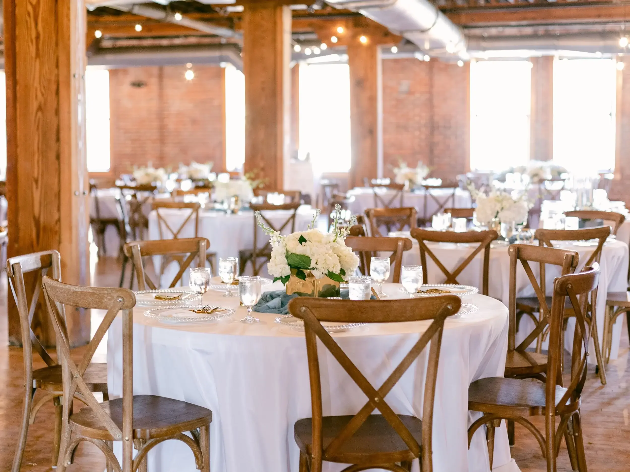 Event space with round tables covered in white tablecloths, decorated with floral centerpieces, set with glassware, and surrounded by wooden chairs, with exposed brick walls and large windows in the background.