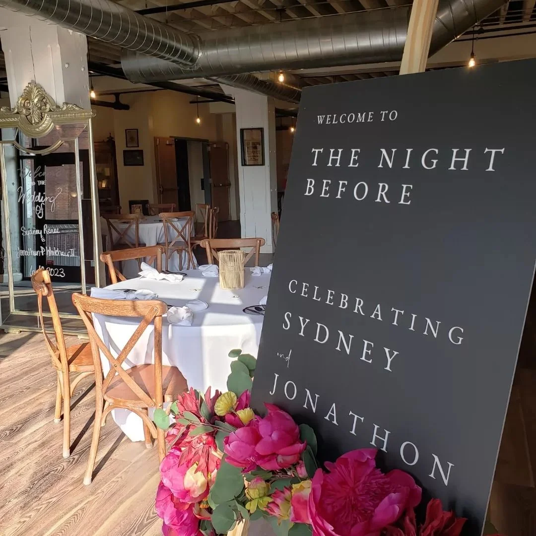 Wedding reception venue with round tables covered in white tablecloths, surrounded by wooden chairs. A black sign with white text welcomes guests to 'The Night Before' celebrating Sydney and Jonathon. Pink flowers are arranged at the base of the sign