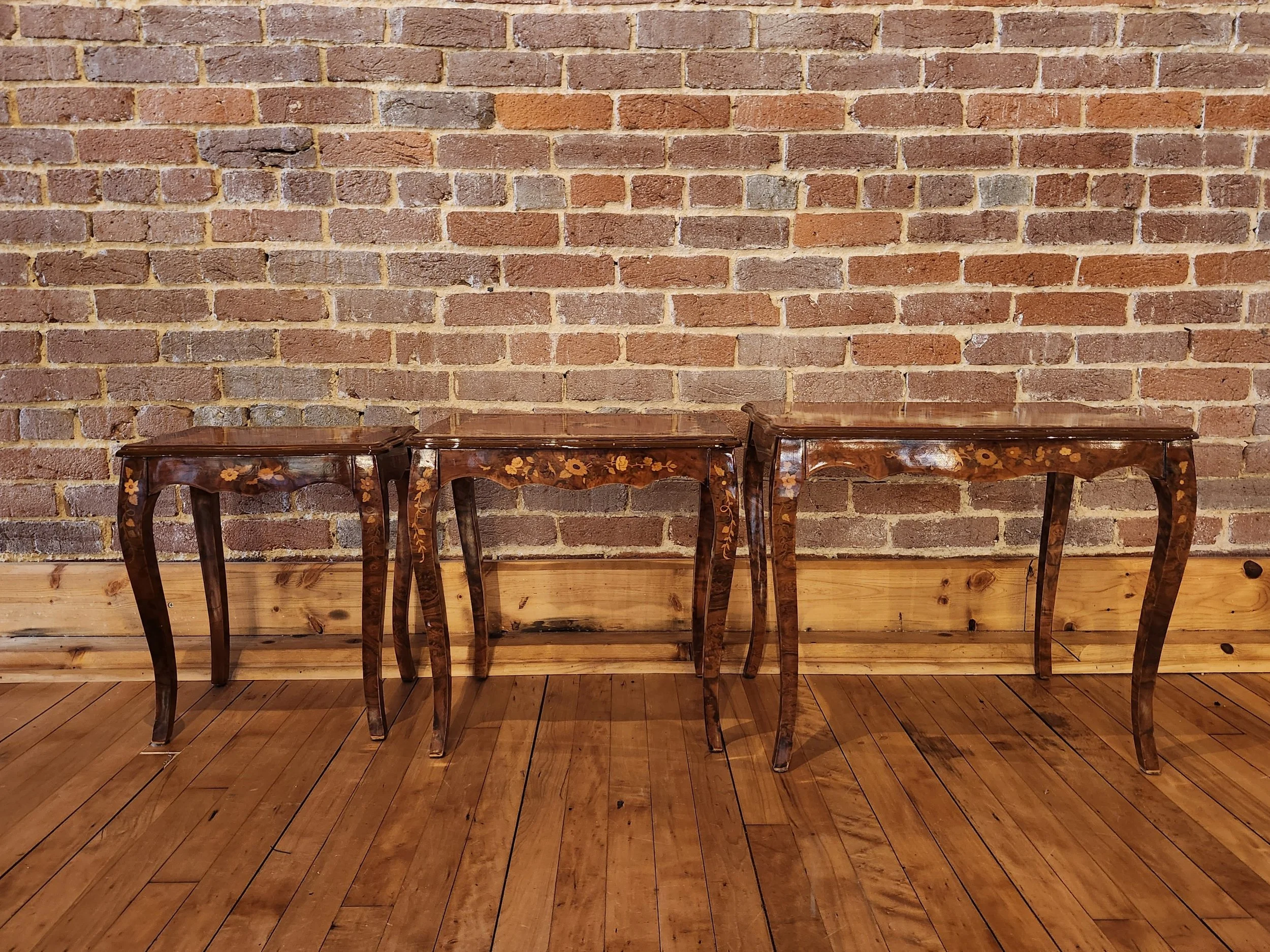 Three vintage wooden side tables with floral inlay design against a brick wall and wooden floor.