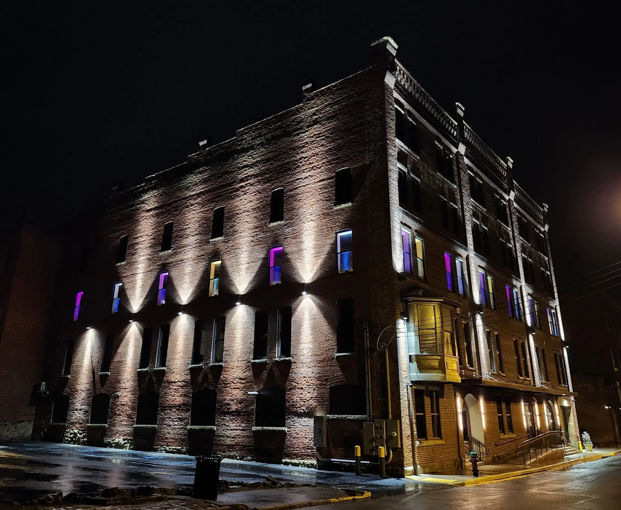 Nighttime view of a multi-story brick building with colorful illuminated windows and exterior lighting highlighting the architecture, wet street in front.
