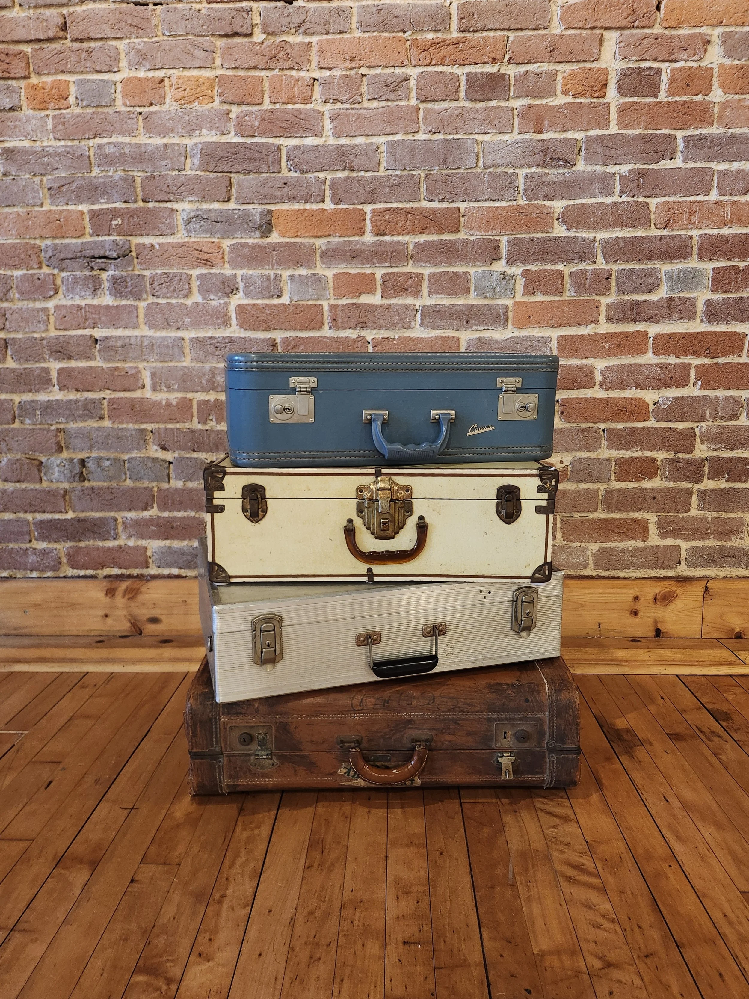 A stack of four vintage suitcases of various sizes and colors against a brick wall, with wooden flooring.