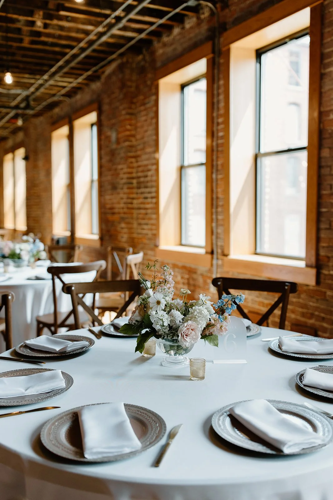 Elegant round banquet table set for an event with white tablecloth, silver chargers, white napkins, gold utensils, floral centerpiece, and table number, in a rustic brick-walled venue with large windows and wooden chairs.