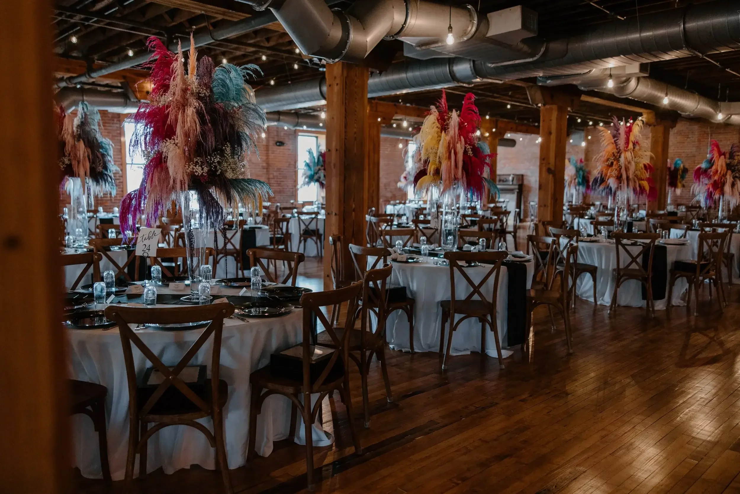 Wedding reception venue with round tables decorated with colorful feather centerpieces, set with glassware and black napkins, in a rustic brick and wood interior.