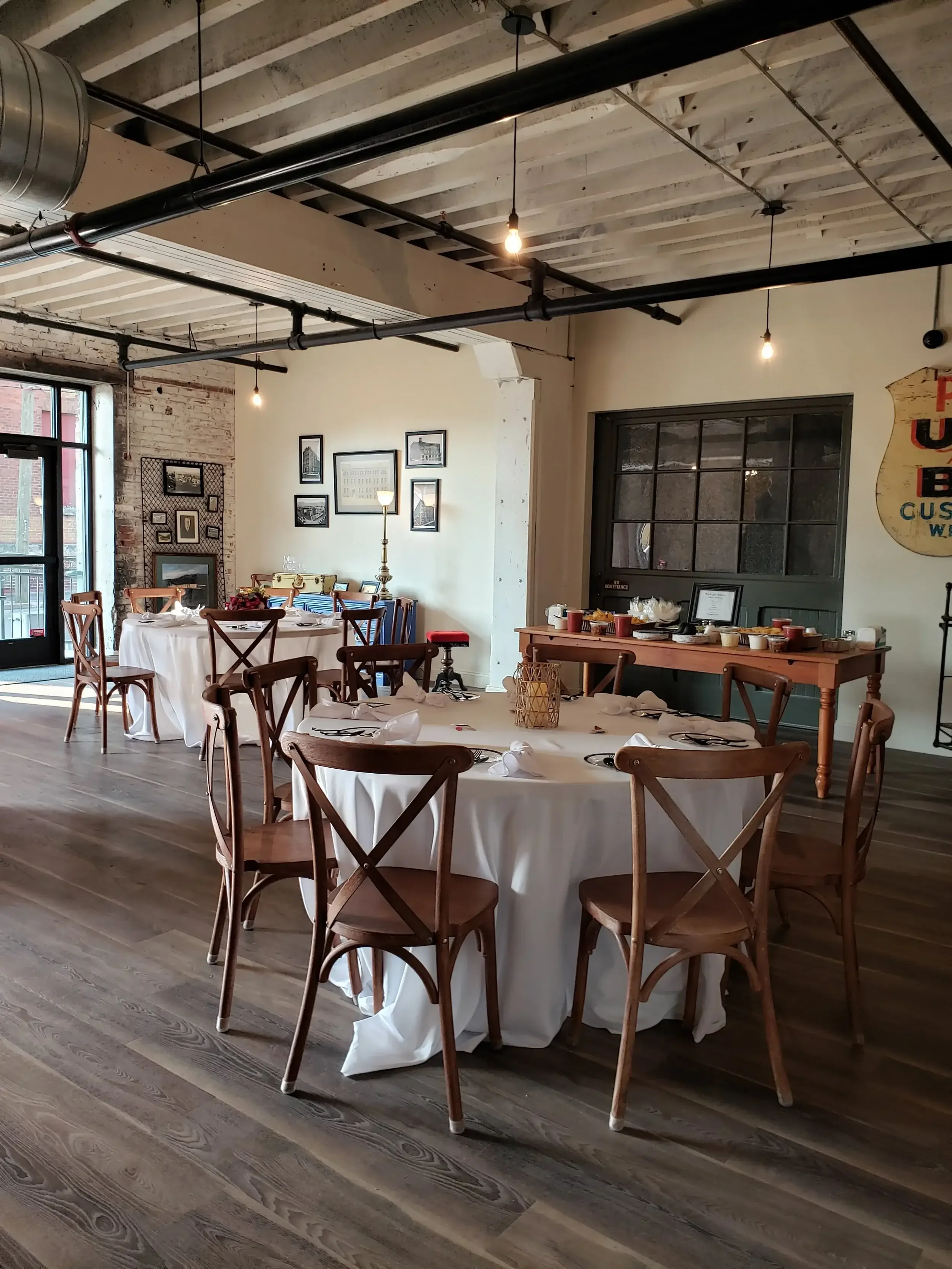 Interior of a restaurant with round tables covered in white tablecloths, set with napkins, utensils, and glasses. Wooden chairs surround the tables. The room has an industrial style with exposed ceiling beams, pipes, and hanging light bulbs. There ar
