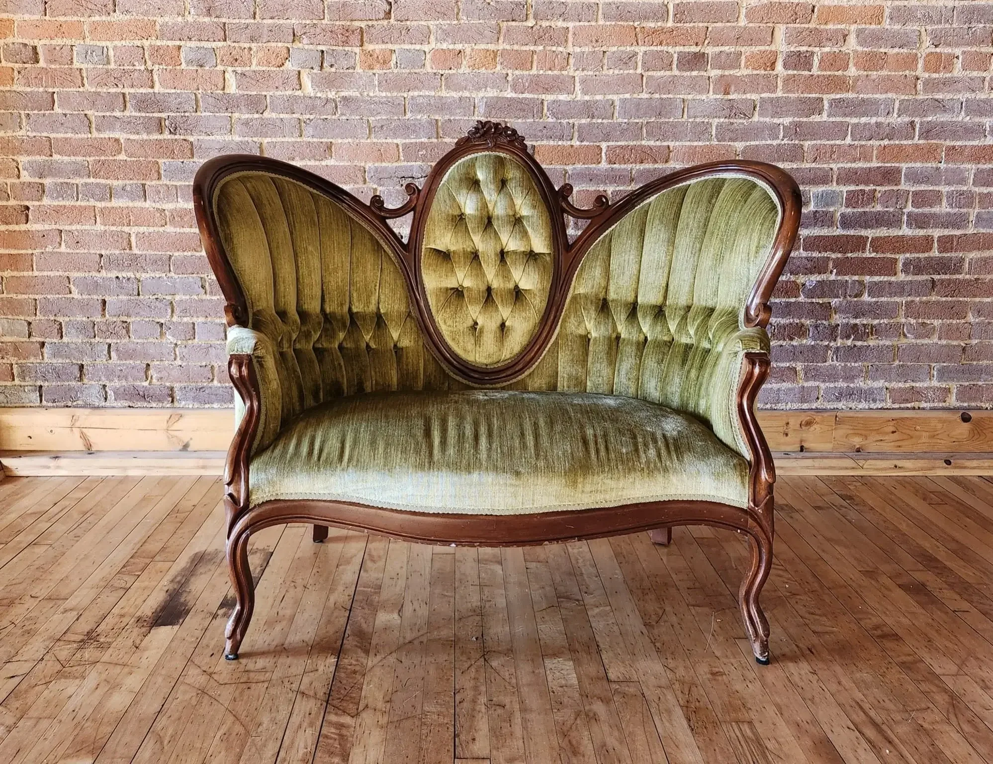 Vintage green velvet settee with carved wooden frame and floral details, positioned against a brick wall on a wooden floor.