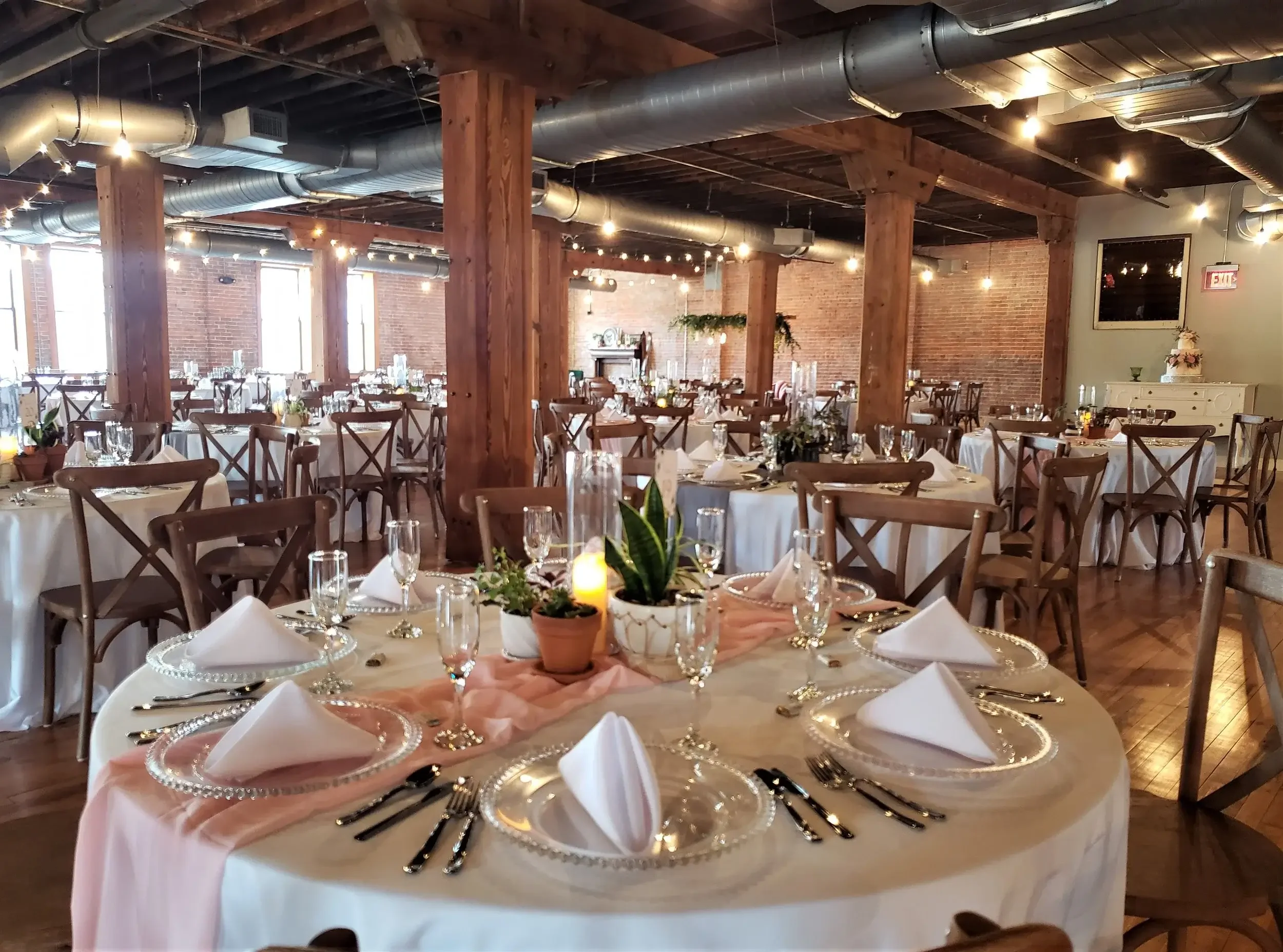 Elegant event space set up for a celebration with round tables covered in white tablecloths, decorated with pink runners, flower centerpieces, candles, and place settings with white napkins folded on clear glass chargers, in a rustic-brick and wooden