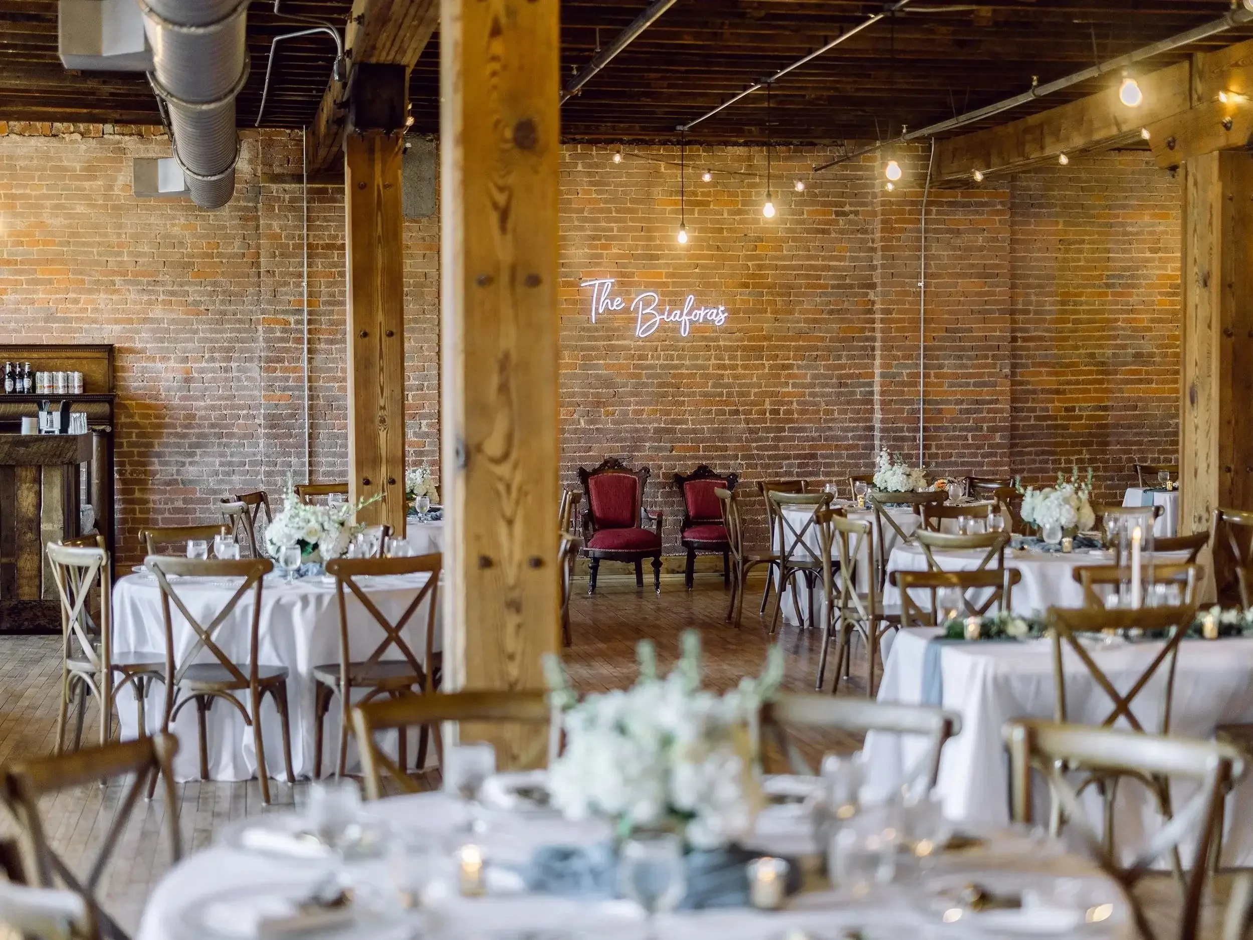 Event space with round tables covered in white tablecloths, decorated with floral centerpieces, and surrounded by wooden chairs, set against a brick wall with a neon sign that reads 'The Bioforas' and string lights hanging from the ceiling.