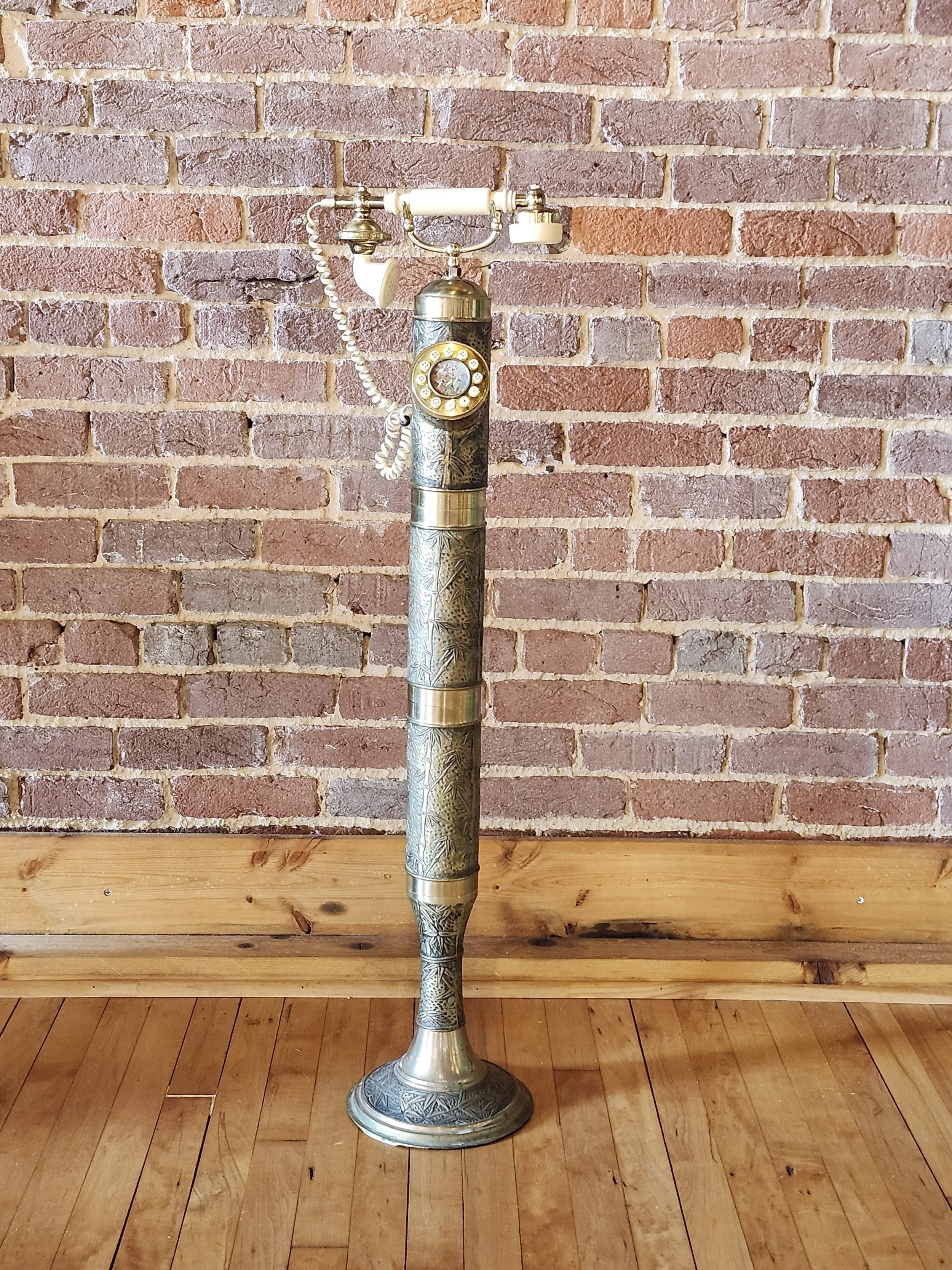 A freestanding vintage-style telephone stand with an ornate metal cover and a rotary phone mounted on top, positioned against a brick wall on a wooden floor.