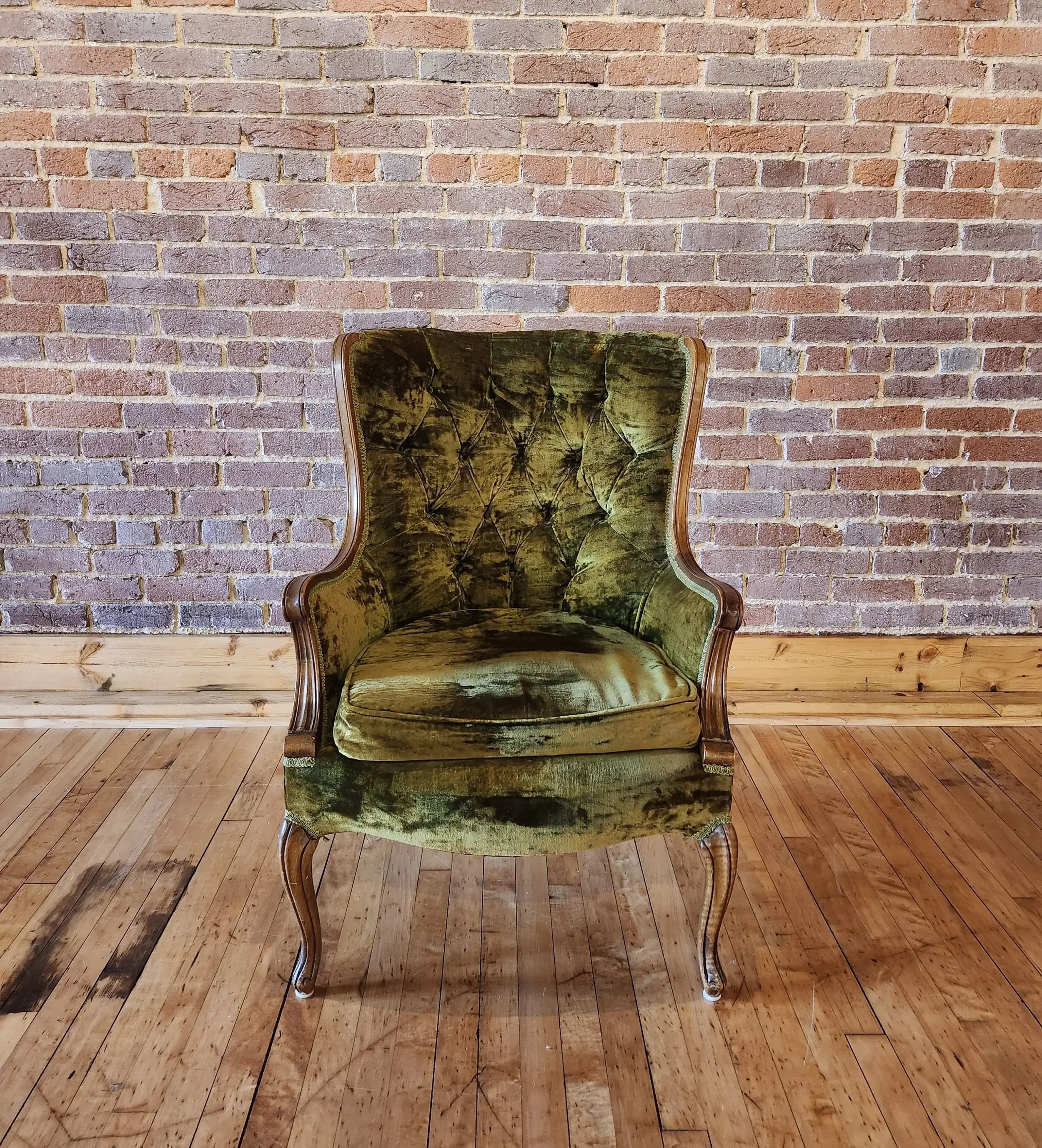 An antique green velvet armchair with wooden frame and curved legs, positioned against a brick wall on a wooden floor.