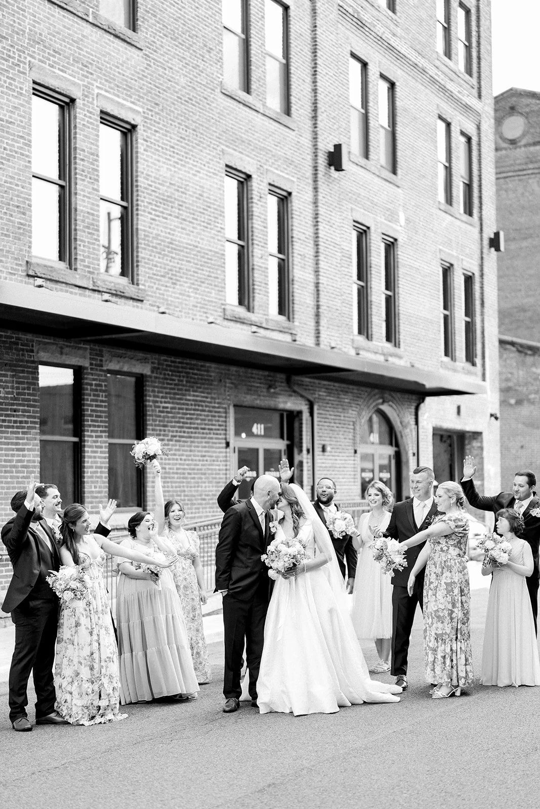 A black and white photo of a wedding celebration outside a brick building. The bride and groom are kissing, surrounded by friends and family who are smiling, waving, and holding flowers.