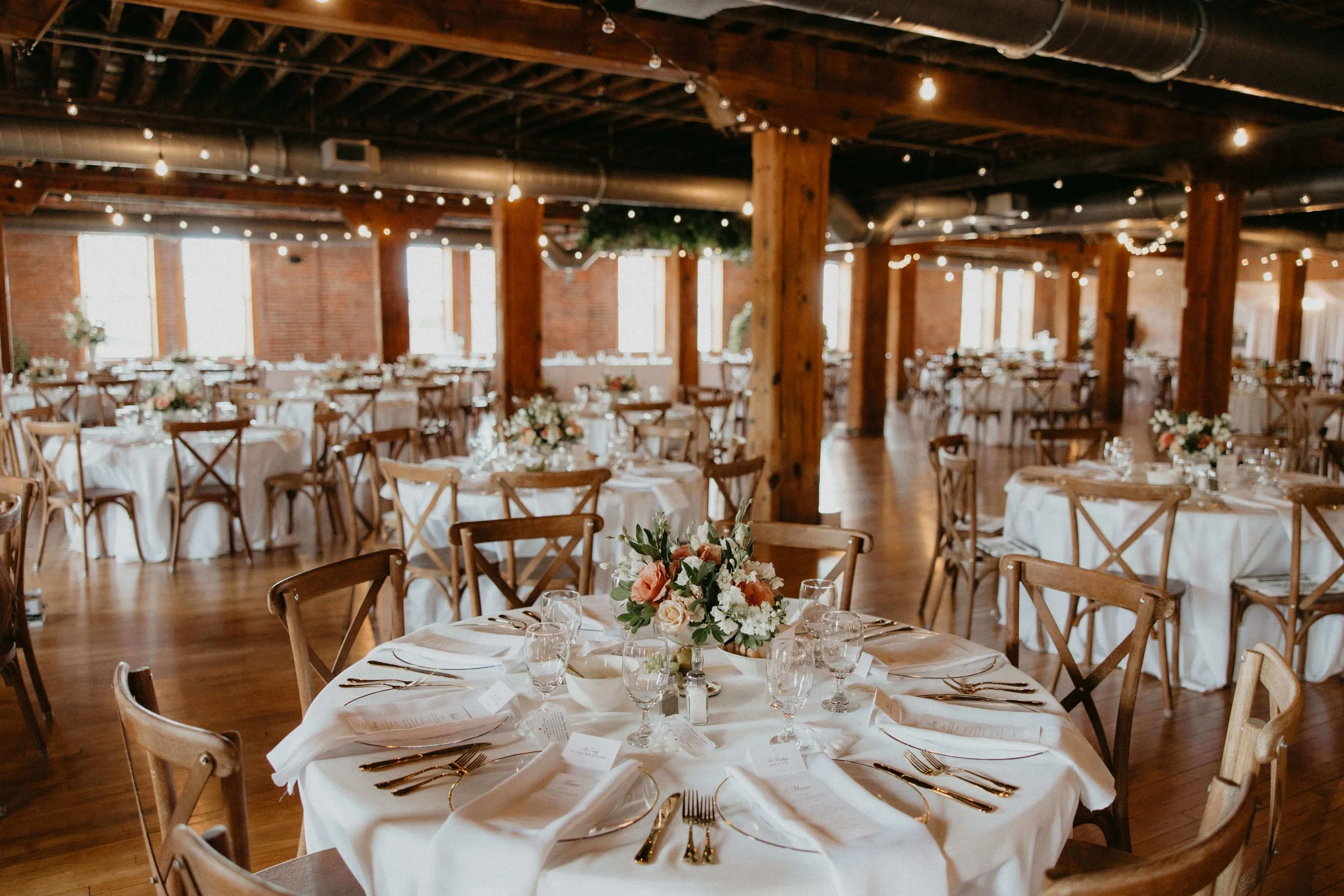 Elegant wedding reception hall with round tables covered in white tablecloths, set with gold utensils, glassware, and white napkins. Each table has a floral centerpiece with pink and white flowers. The hall features exposed brick walls, wooden beams,