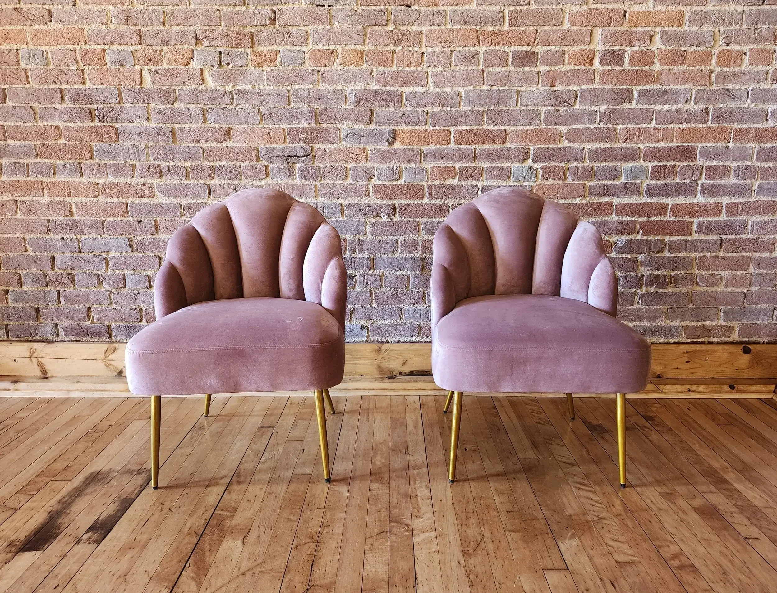 Two pink velvet accent chairs with gold legs are positioned side by side against an exposed brick wall in a room with wooden flooring.