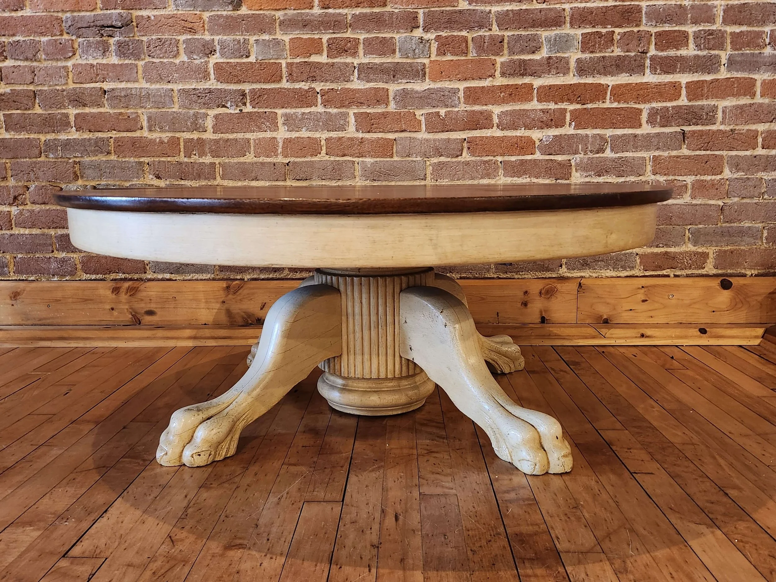 Wooden table with a carved pedestal base, set against a brick wall, on a wooden floor.