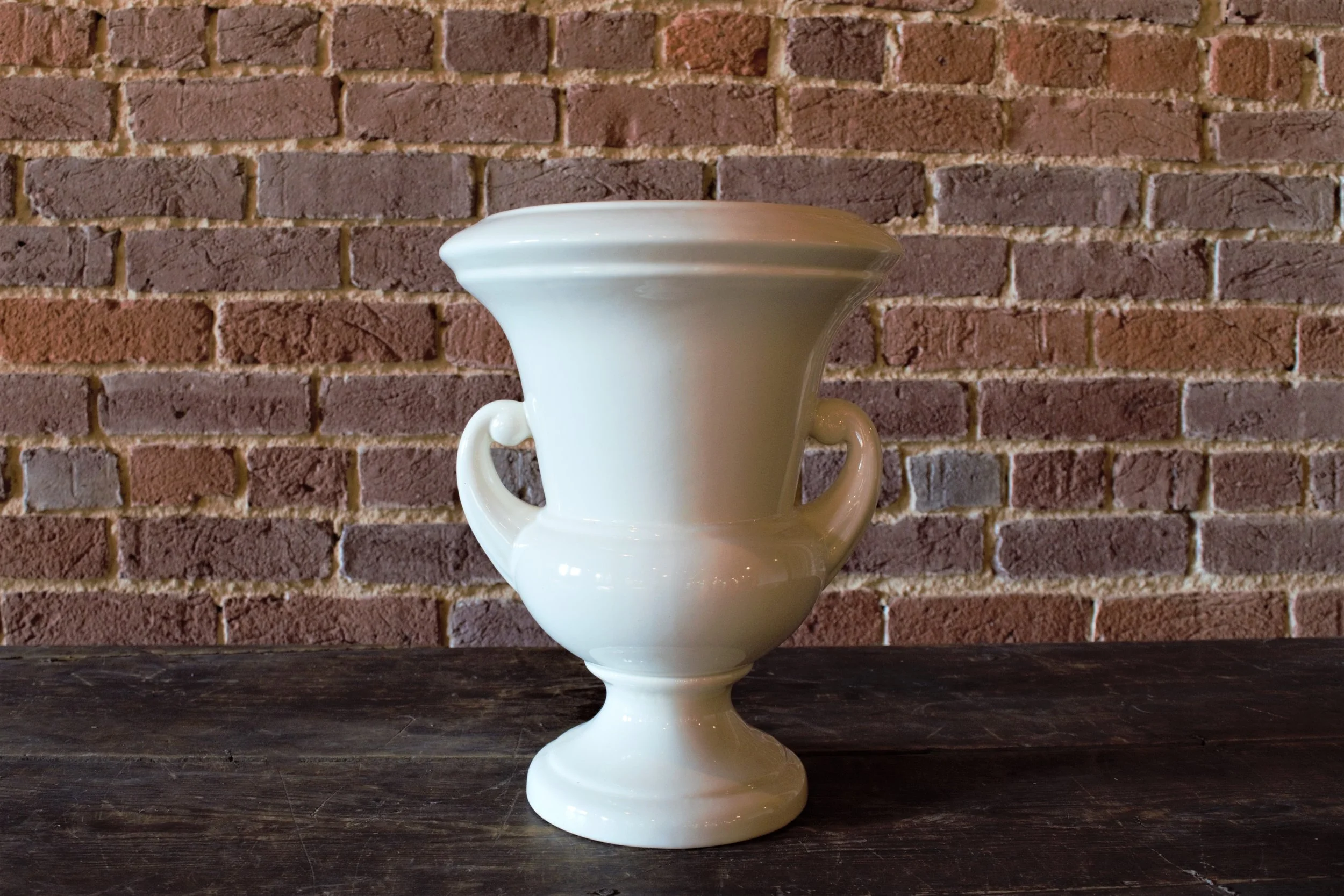 A white ceramic vase with two handles, placed on a dark wooden surface against a brick wall background.