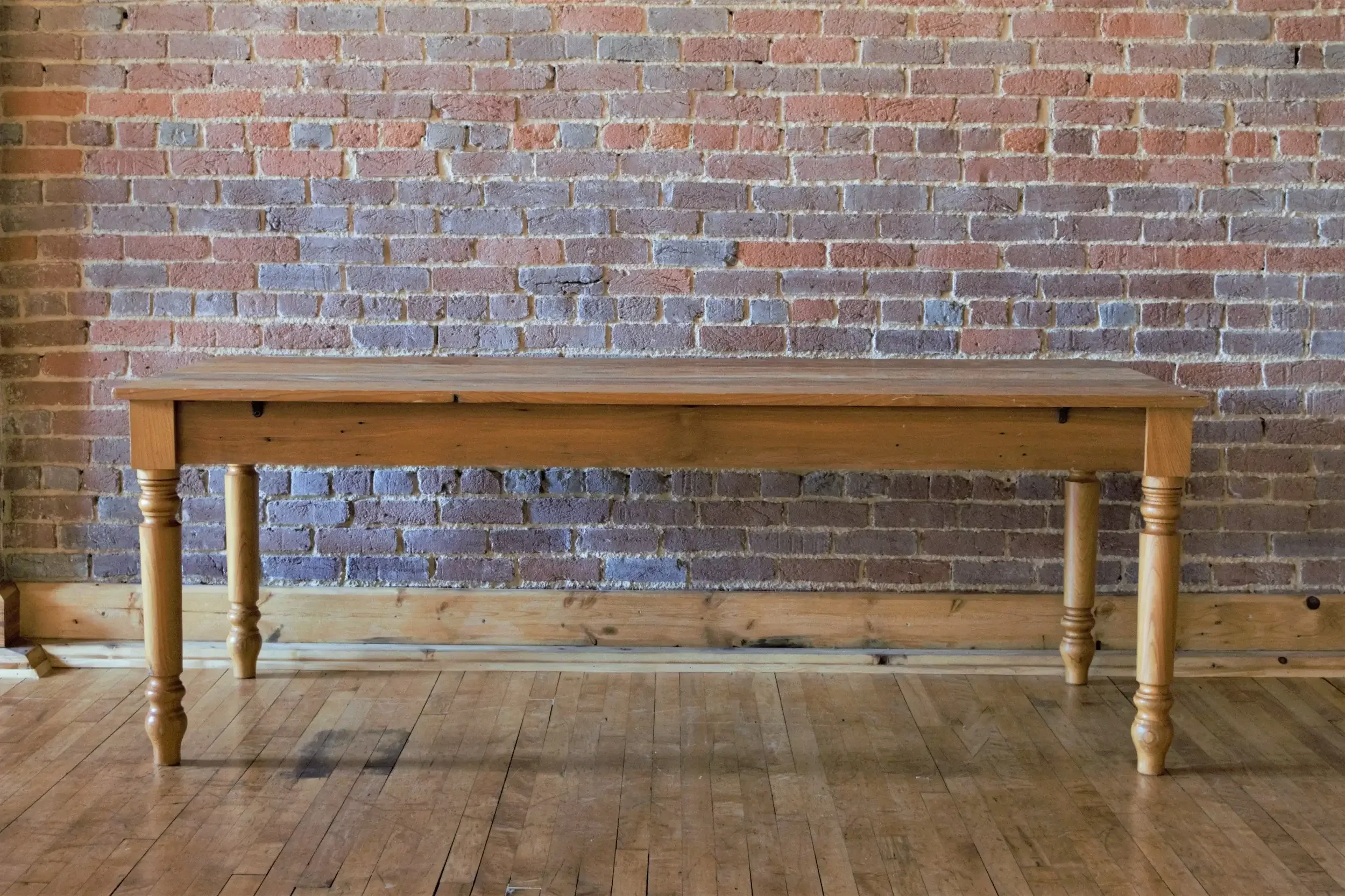 A wooden table with turned legs placed against an exposed brick wall, sitting on a hardwood floor.