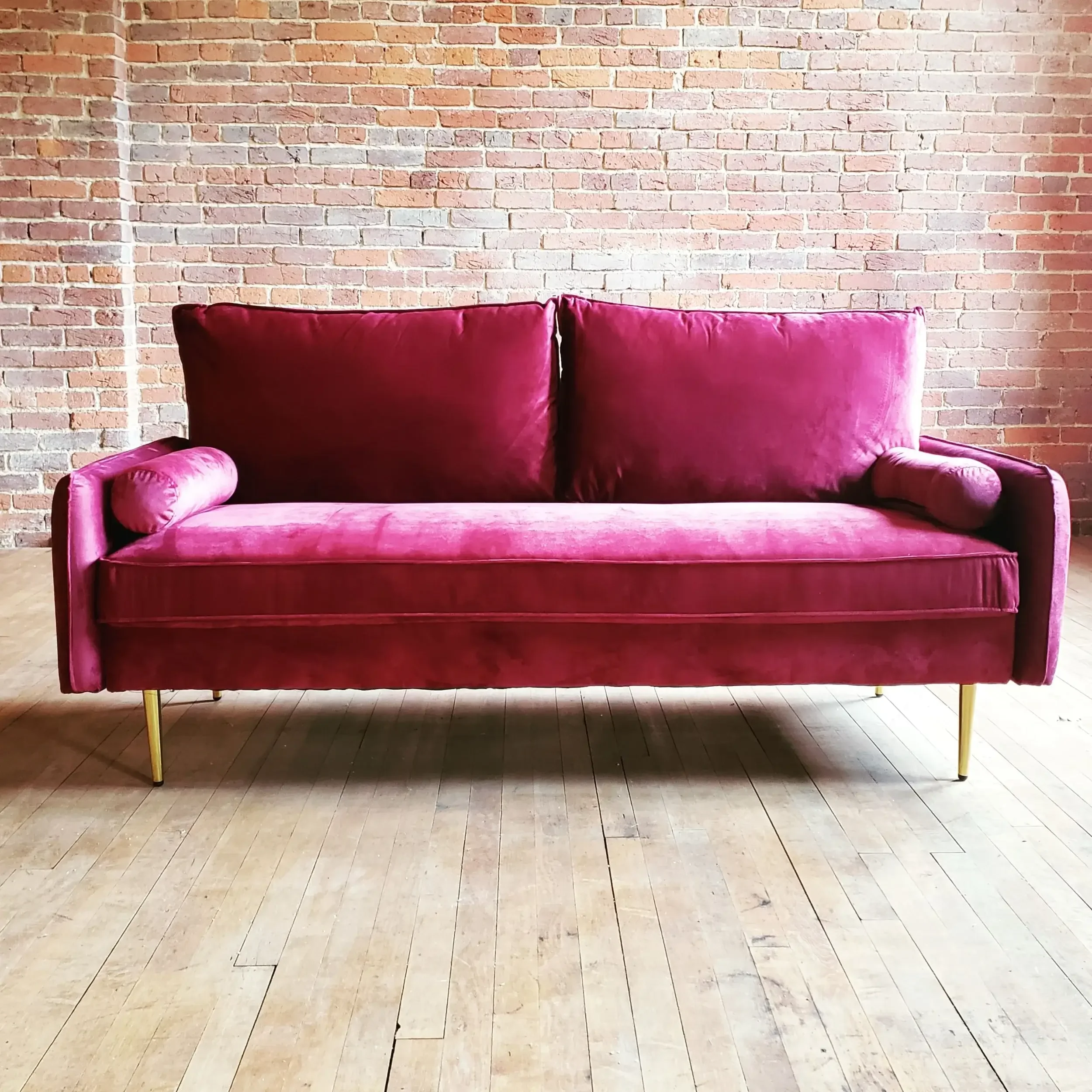 Red velvet sofa with gold-toned legs against a brick wall and wooden floor