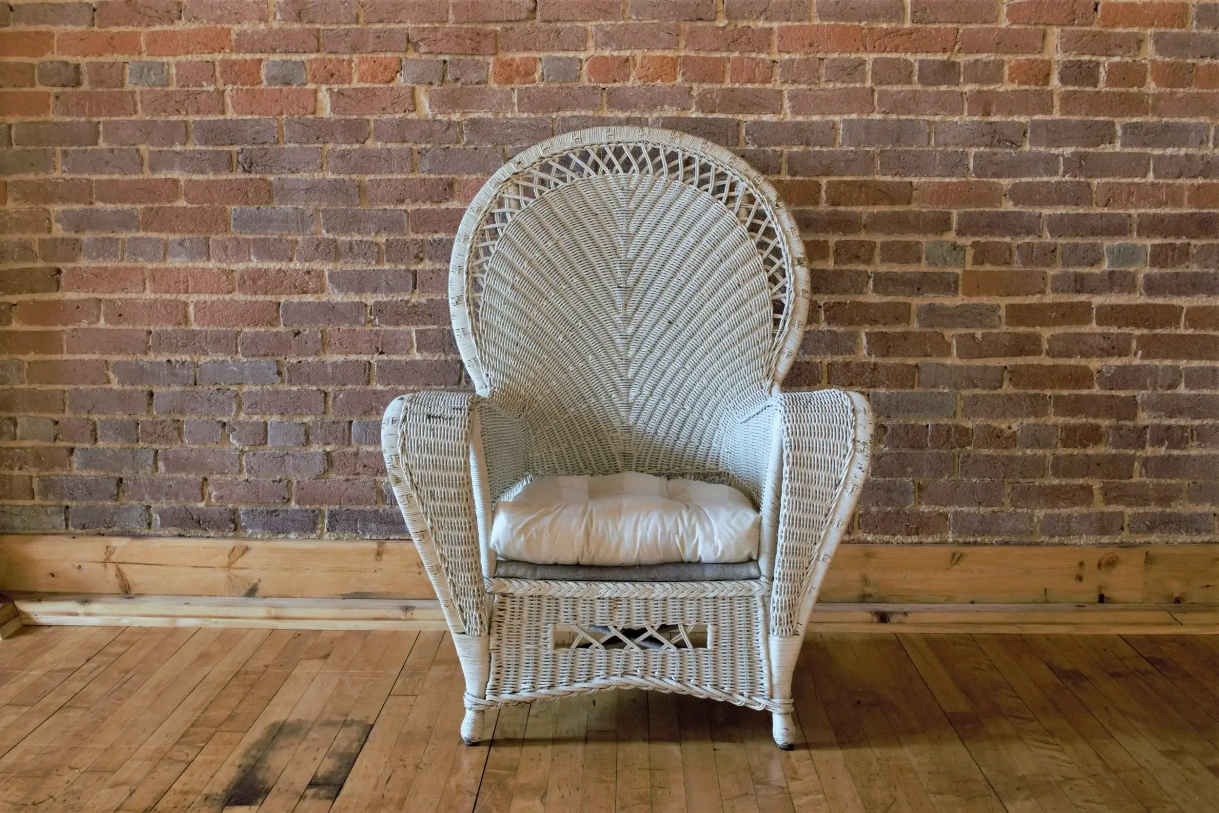 Wicker high-back chair with a light-colored cushion, positioned against a brick wall and placed on a wooden floor.