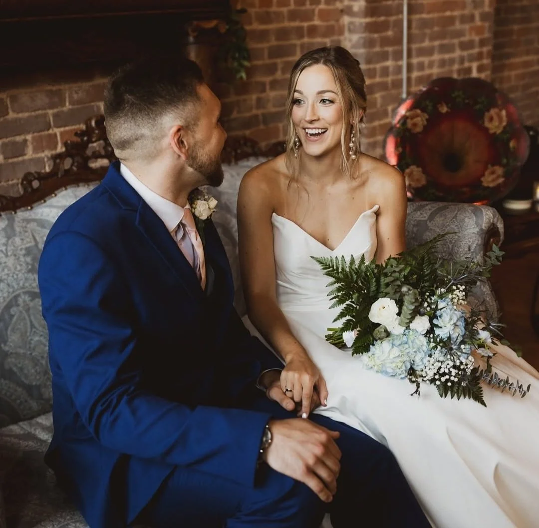 A bride and groom sitting on a vintage sofa, holding hands, smiling at each other, in an indoor setting with brick walls, during their wedding reception.