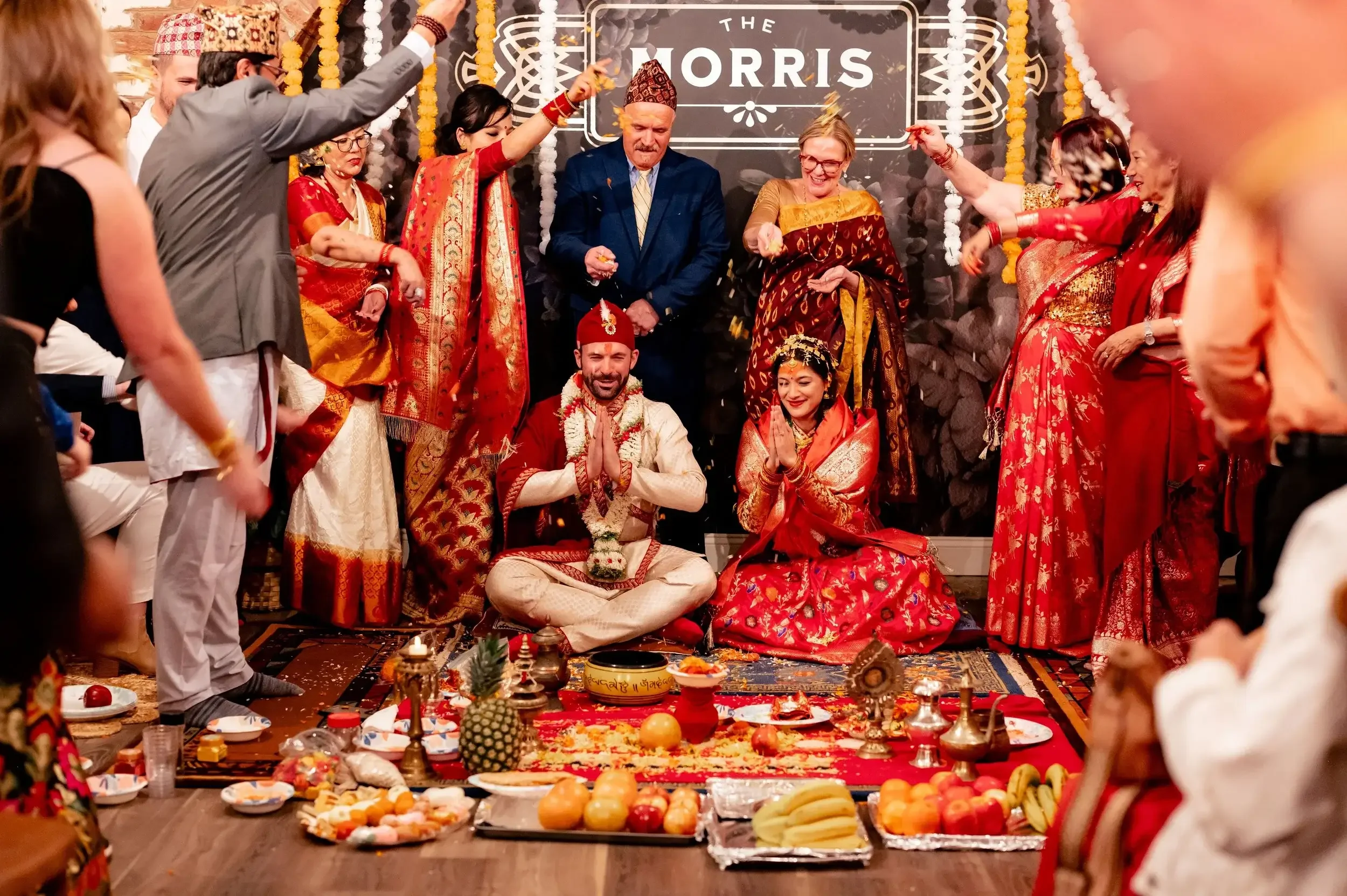 A traditional Hindu wedding ceremony with the bride and groom sitting on the floor, surrounded by family and friends dressed in colorful clothing. The couple is in front of a decorated altar with offerings, fruits, and flowers, while the guests parti