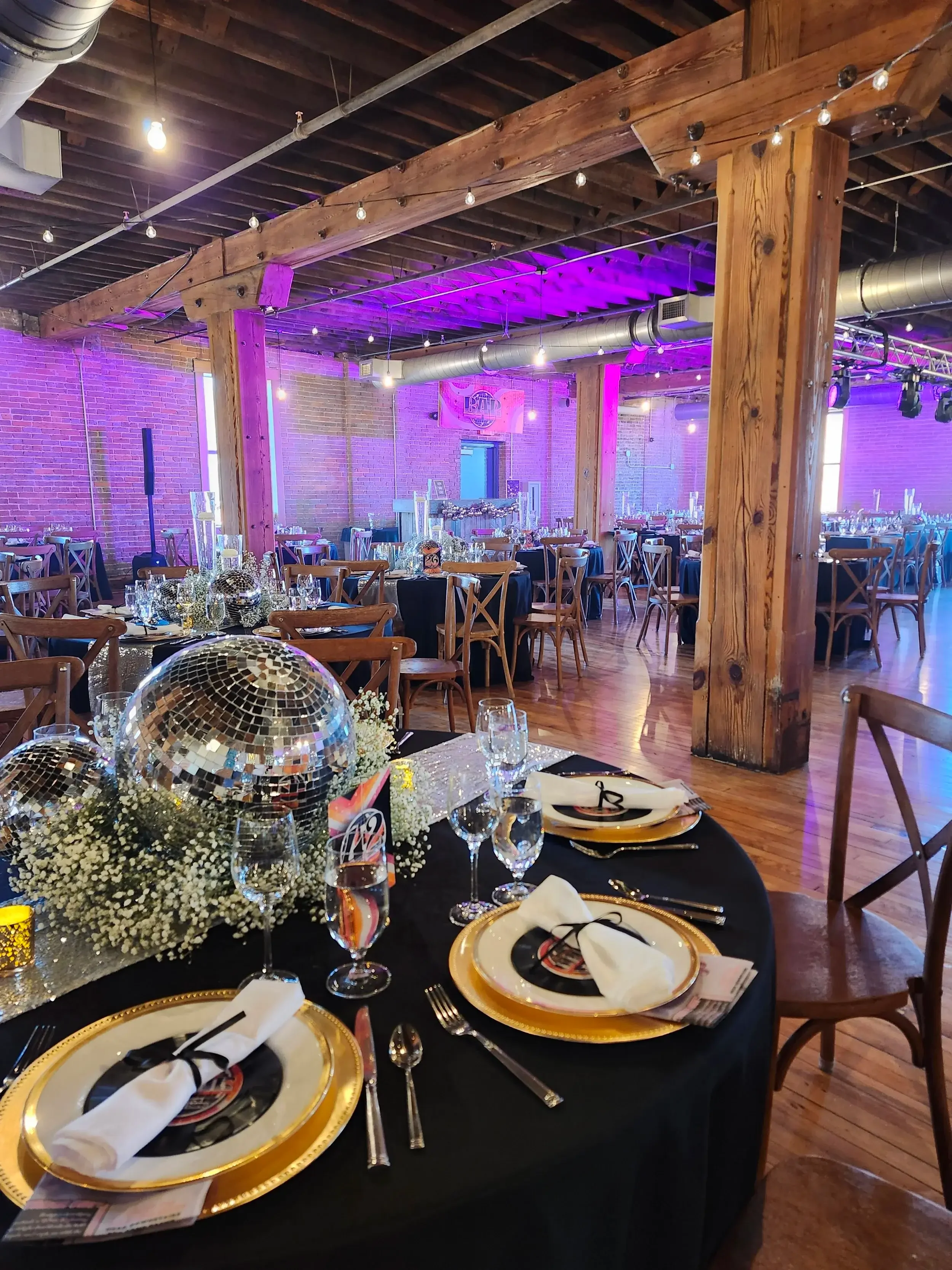 Decorated banquet hall with round tables covered in black tablecloths, gold and white plates, glasses, and centerpieces made of disco balls and white flowers. The hall has wooden beams, exposed brick walls, and purple lighting.
