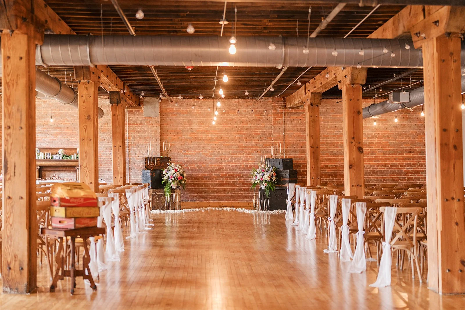 Empty wedding ceremony venue with wooden chairs decorated with white fabric, floral arrangements at the front and brick walls.