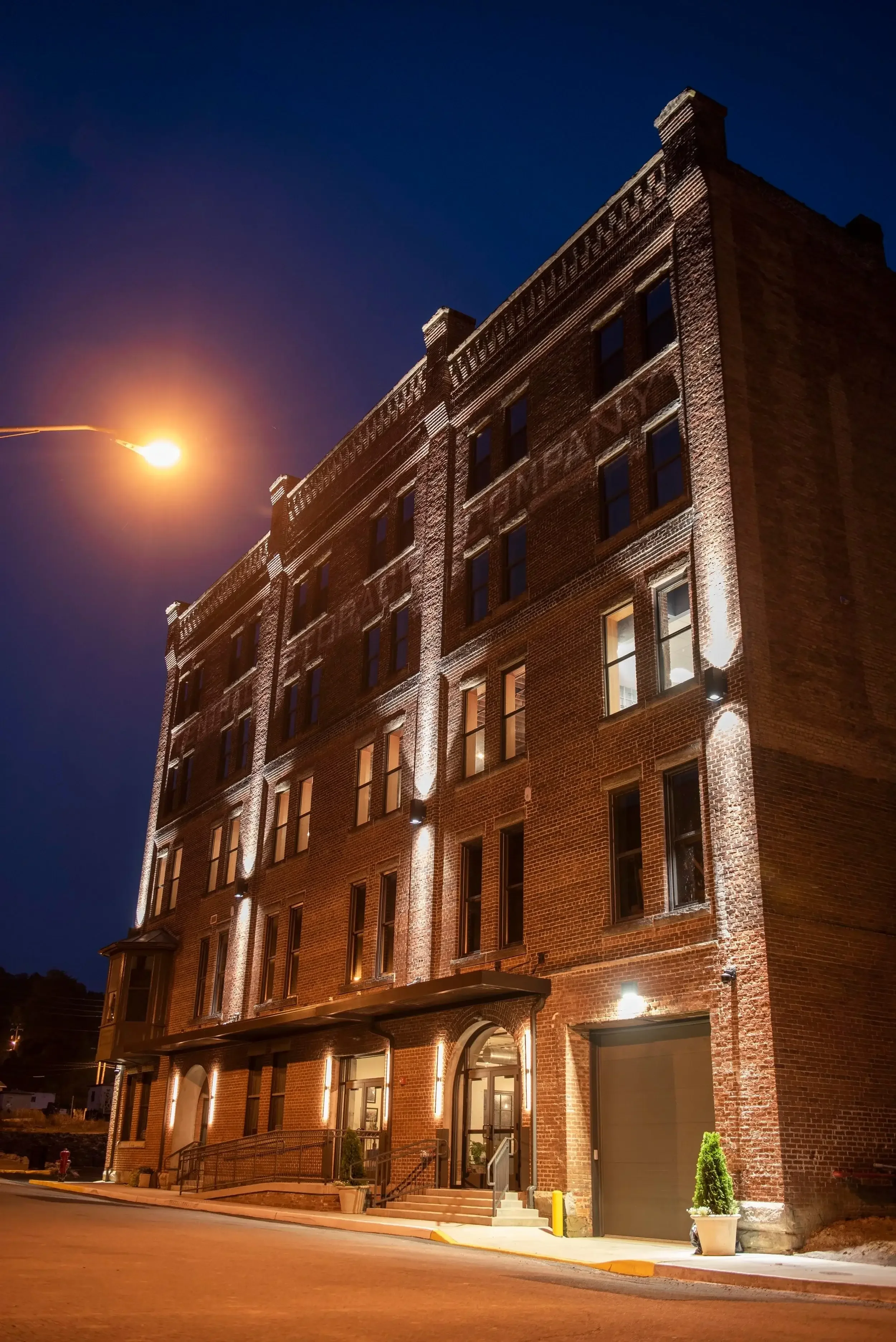 A tall brick building illuminated at night with exterior lighting accents, a wheelchair accessible ramp, and potted plants near the entrance.