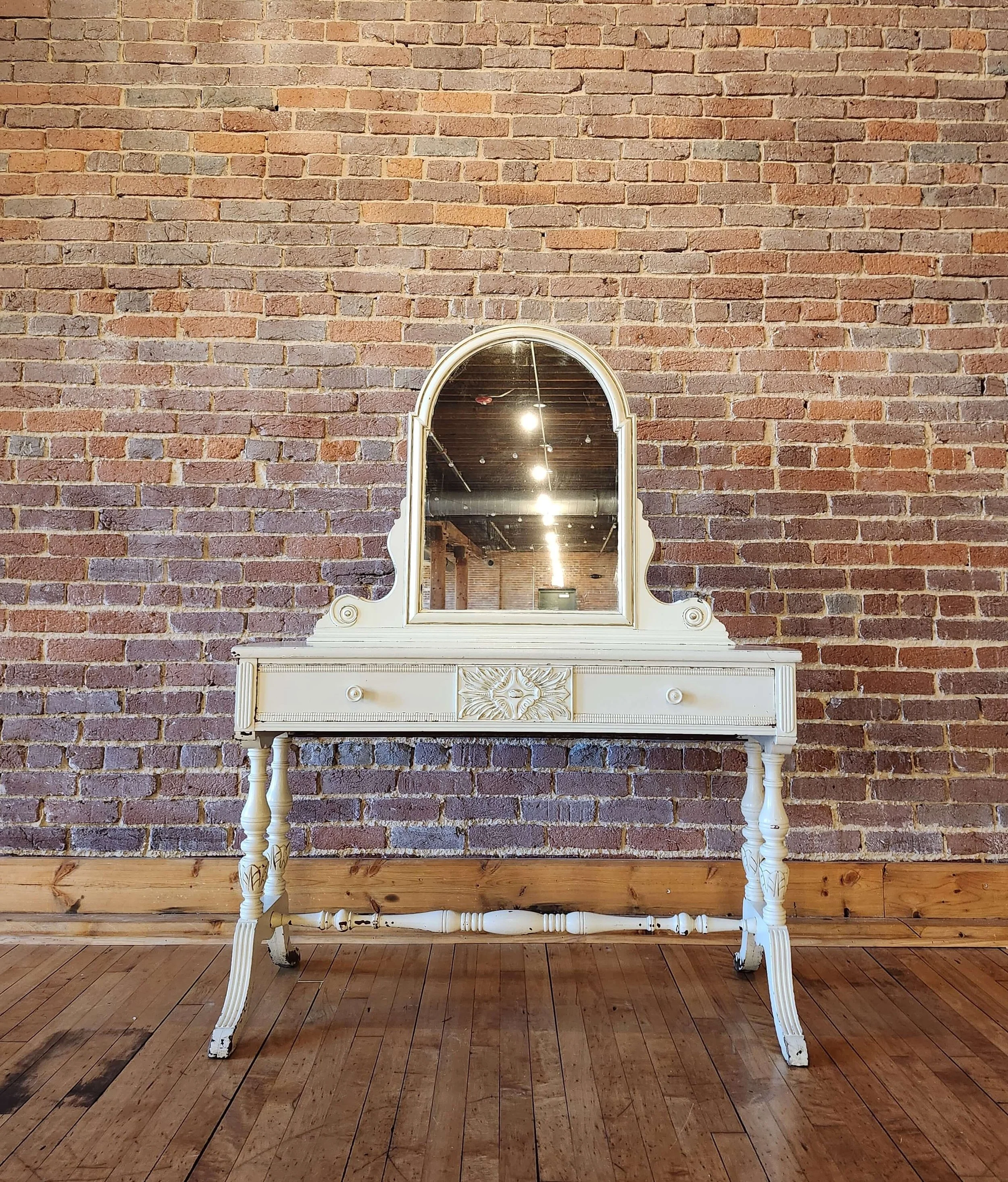 A vintage white vanity table with a mirror, ornate carved details, and turned legs, set against a red brick wall and wooden floor.