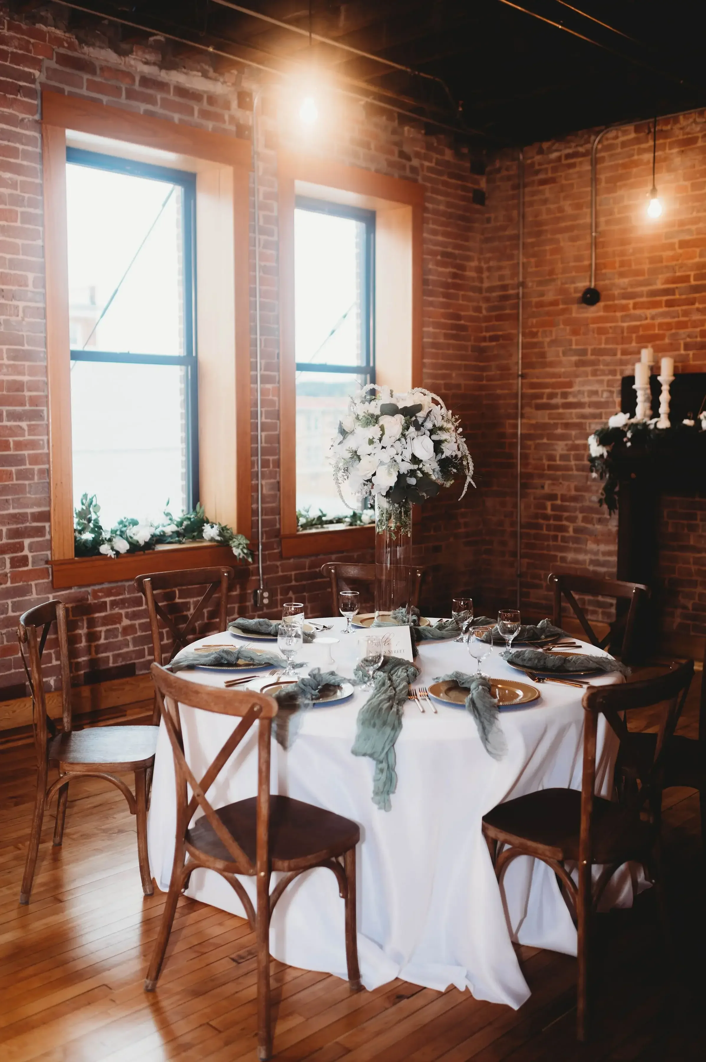 Elegant round dining table with a white tablecloth, surrounded by six wooden chairs, set for a special occasion with napkins, glasses, and plates, and a tall floral centerpiece in a clear vase, in a brick-walled room with large windows letting in nat
