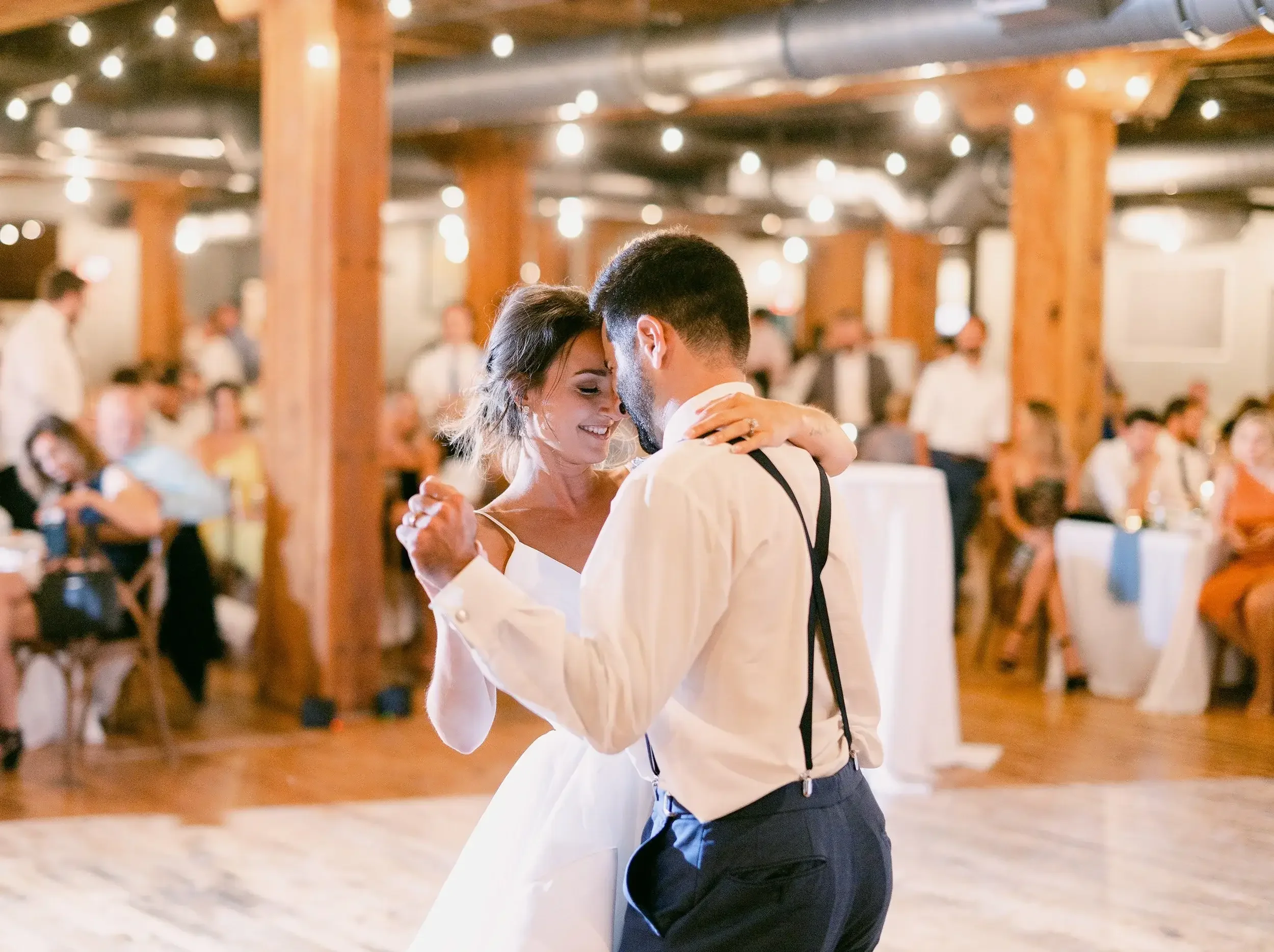 A bride and groom sharing their first dance at their wedding reception in a rustic venue decorated with string lights, with guests seated at tables in the background.