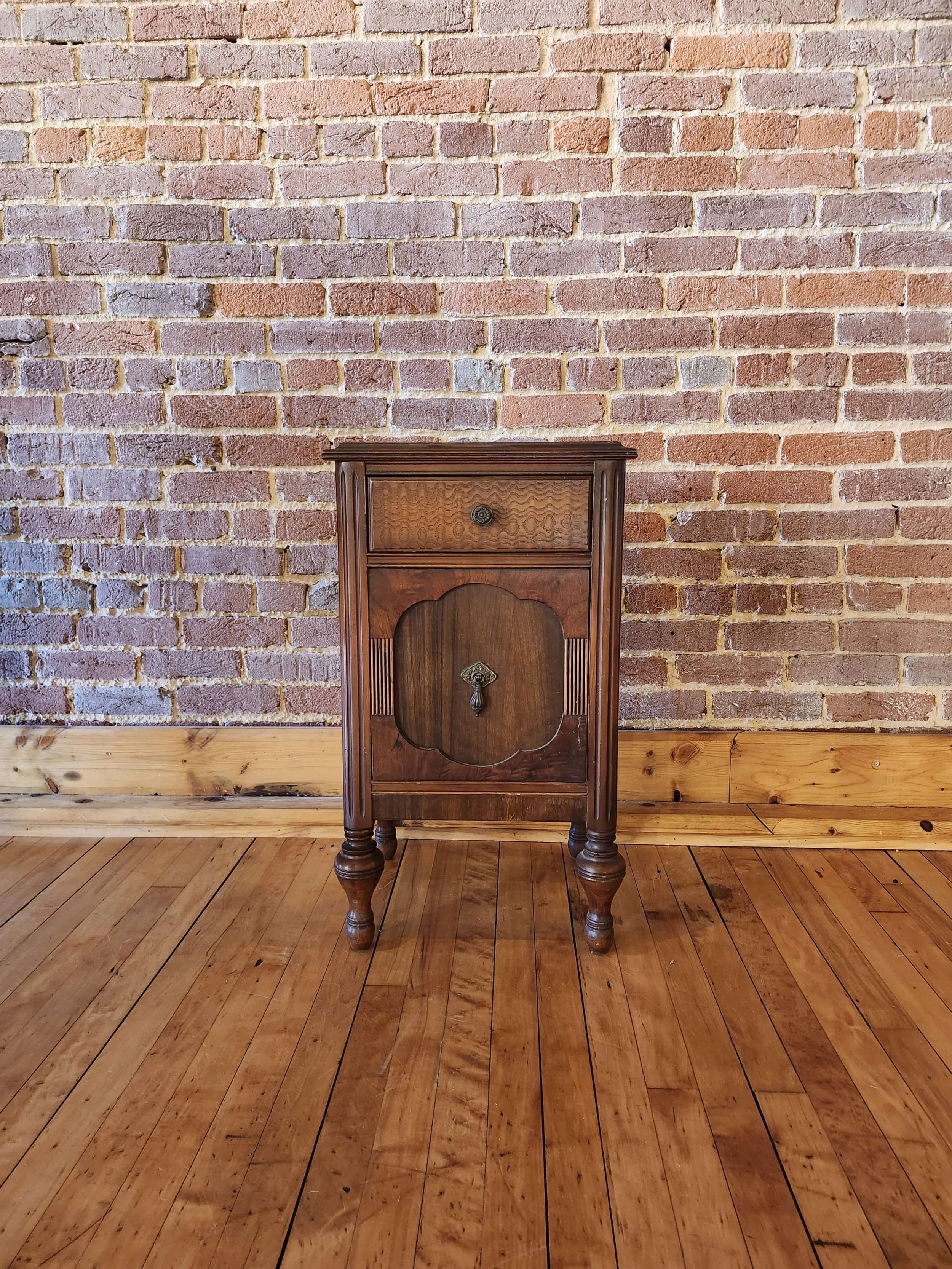 A small, vintage wooden cabinet with a drawer on top and a door below, standing on four turned legs, set against a brick wall with wooden flooring.