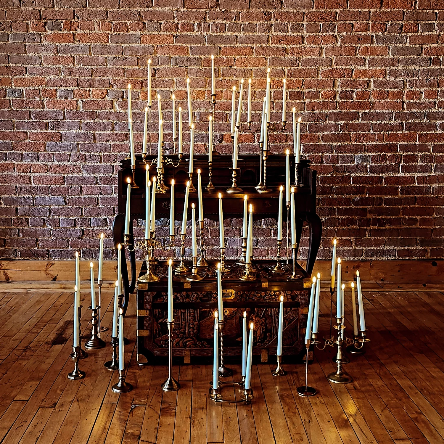 A collection of unlit and lit candles on candle holders placed on and around a wooden chest in front of a brick wall.
