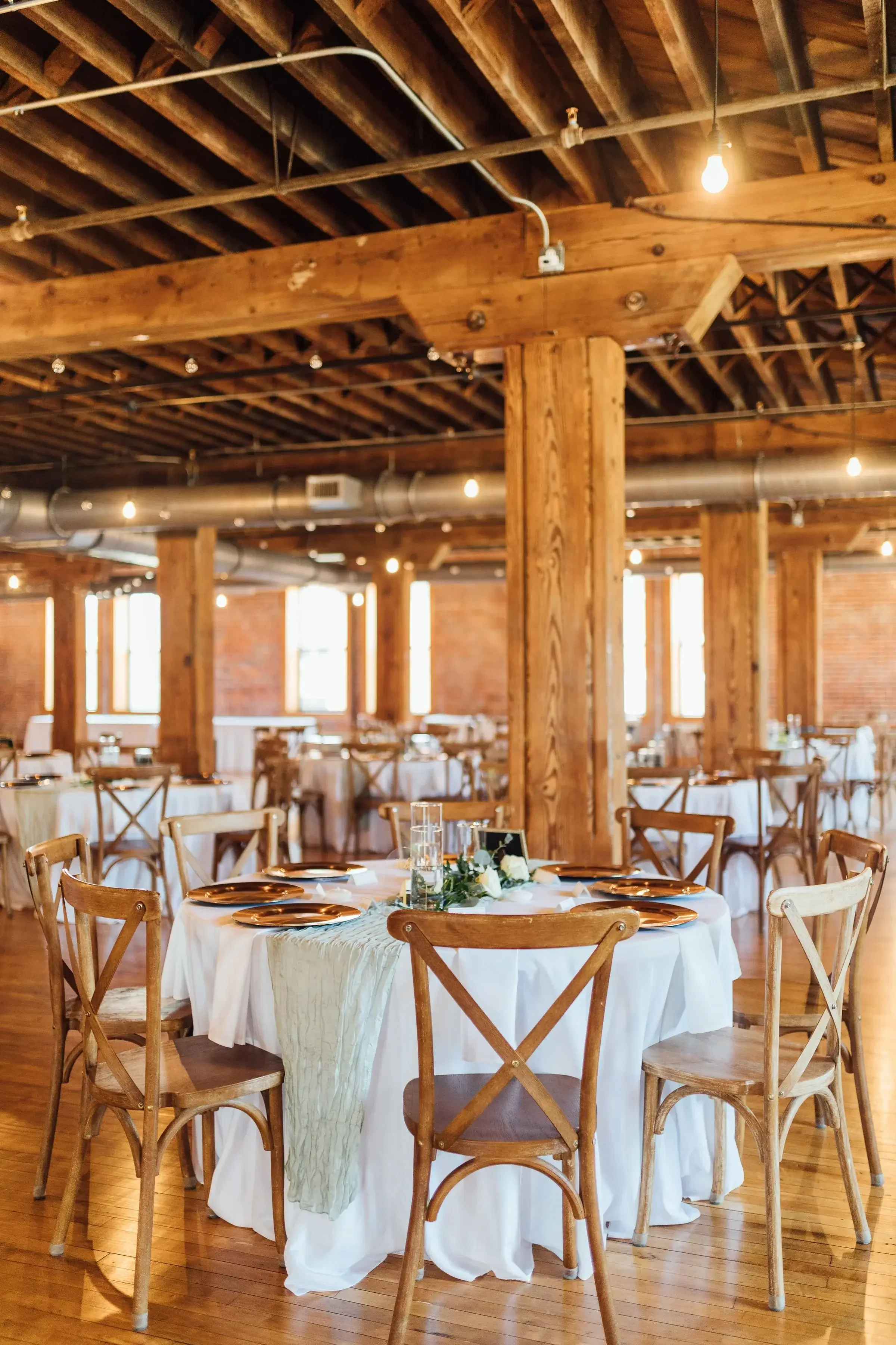 Wedding reception setup with round tables, wooden chairs, and floral centerpieces in a rustic venue with exposed wooden beams and brick walls.