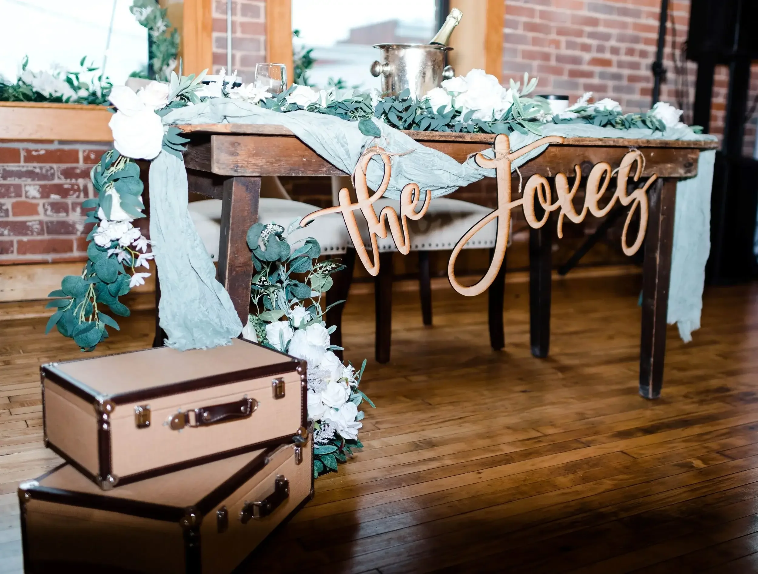 Decorative wooden table with a "the love" sign, white flowers, and green foliage garland, set against a brick wall interior, with stacked vintage suitcases in the foreground.