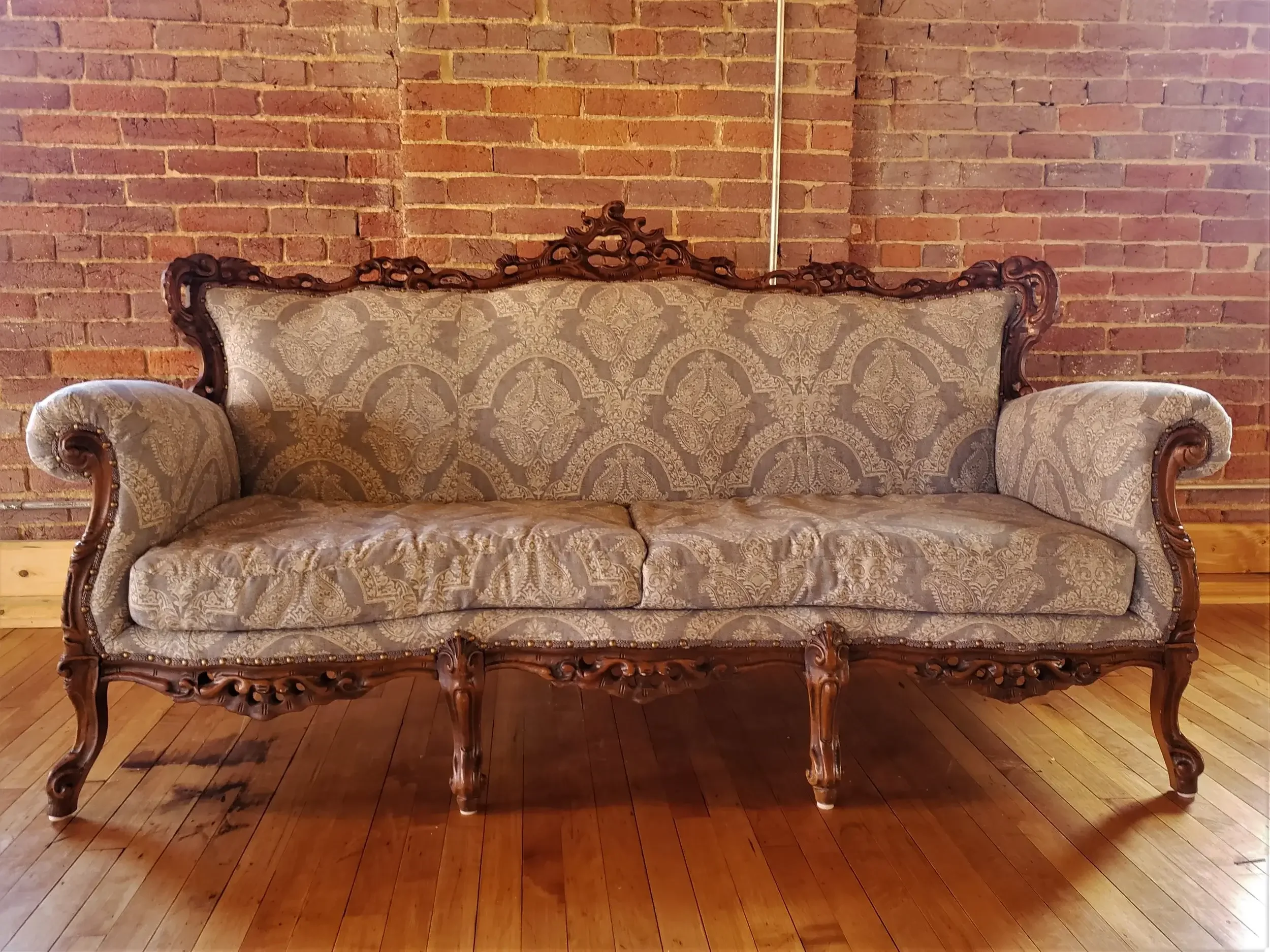 An antique ornate sofa with carved wooden frame and patterned fabric upholstery, placed against a brick wall on a wooden floor.