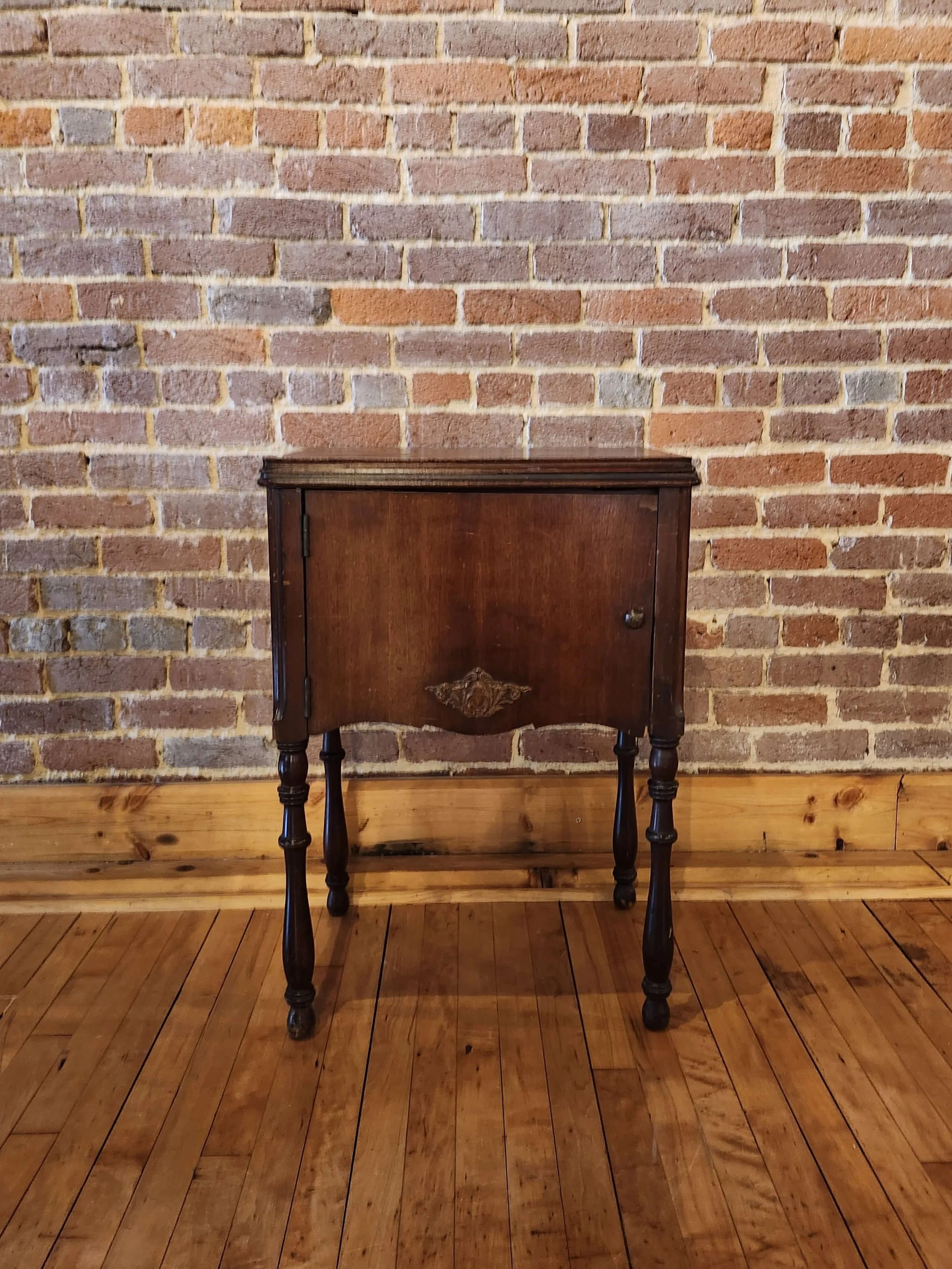 An antique wooden cabinet with a carved decorative element on the front, standing on four turned legs, positioned against a brick wall on a wooden floor.
