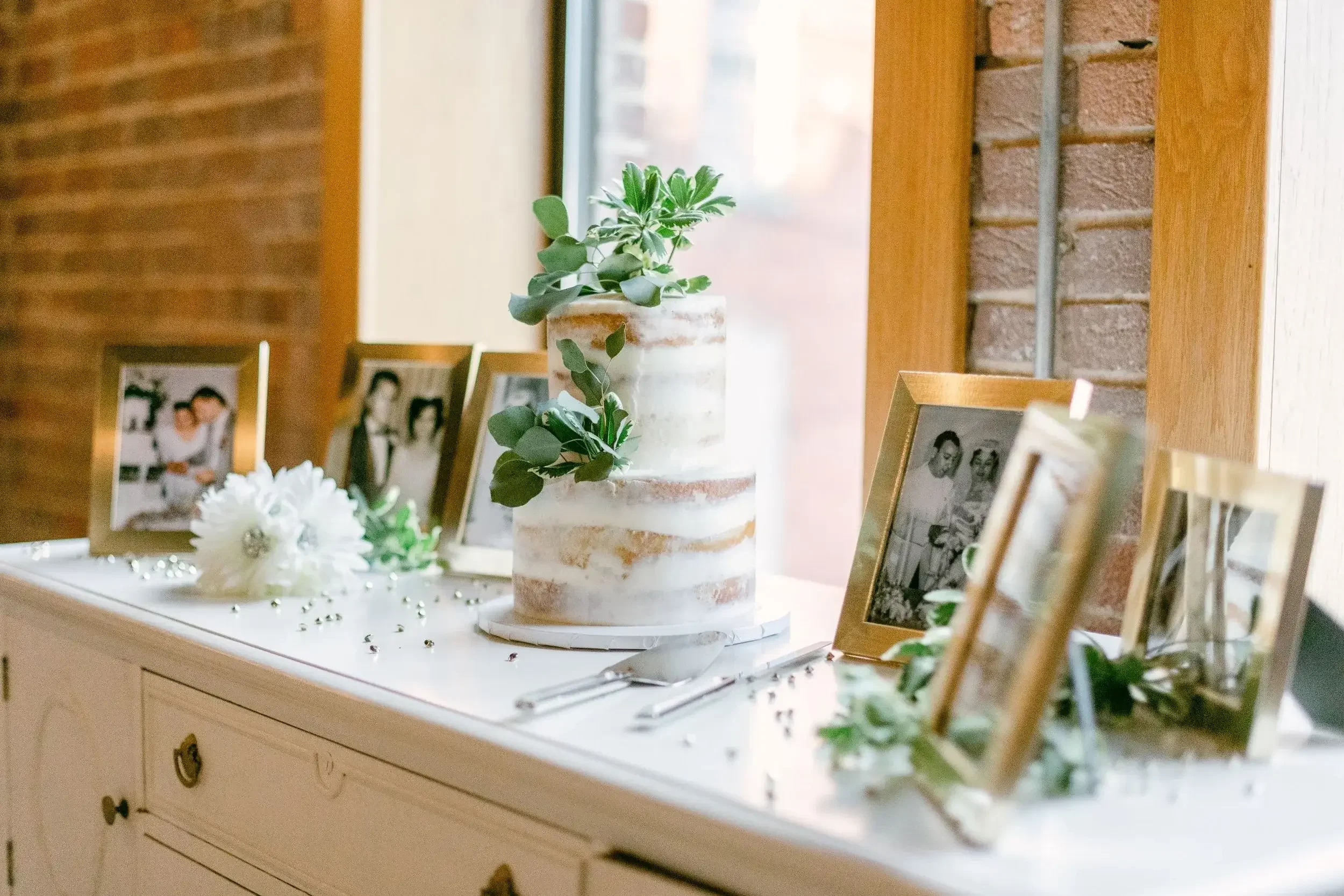 A rustic naked wedding cake with green foliage on top, displayed on a white sideboard alongside framed black and white photographs, white flowers, and scattered silver beads.