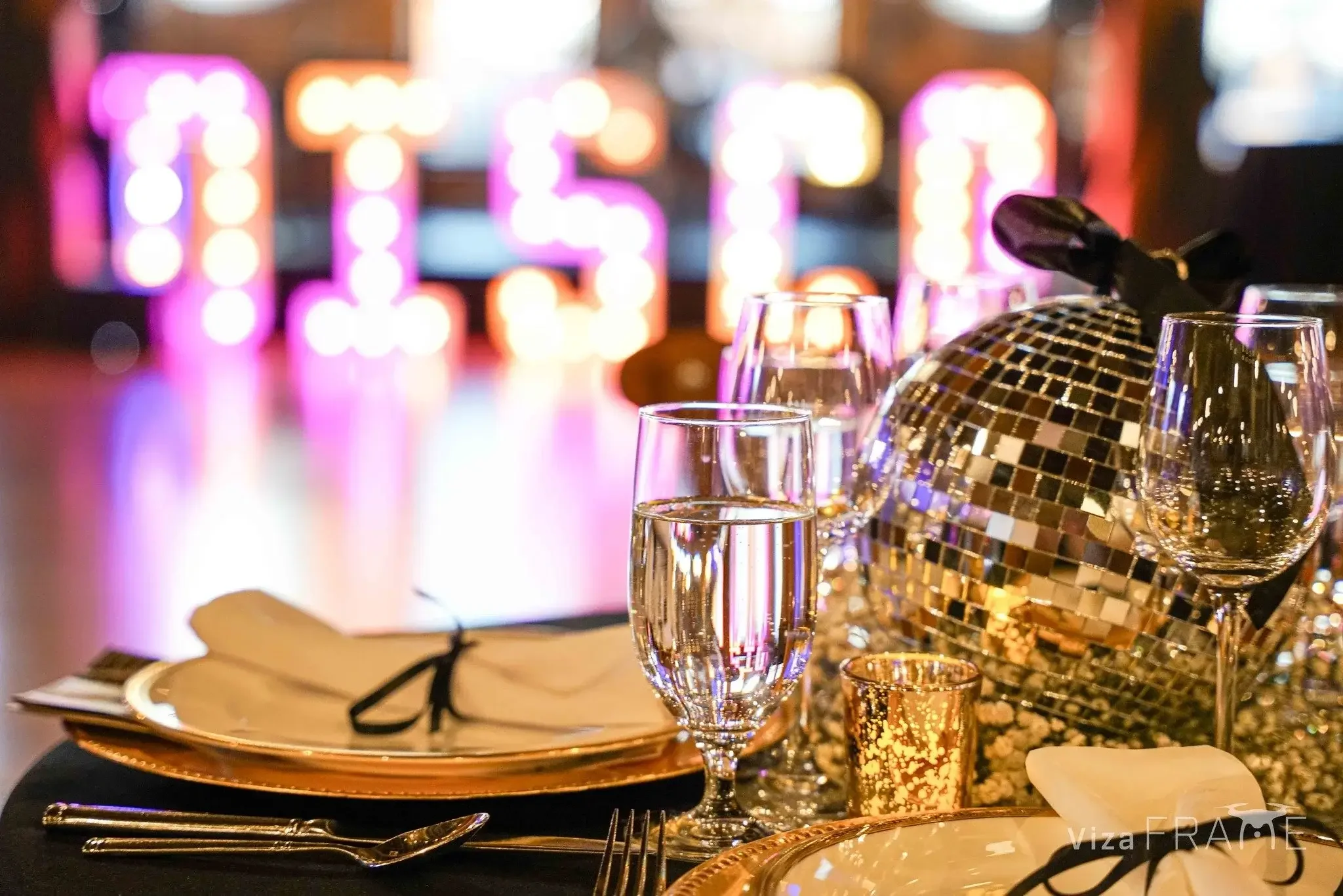 Elegant table setting with gold-rimmed plates, wine glasses, and a gold disco ball centerpiece, set against a colorful, blurred background with pink and purple lights.