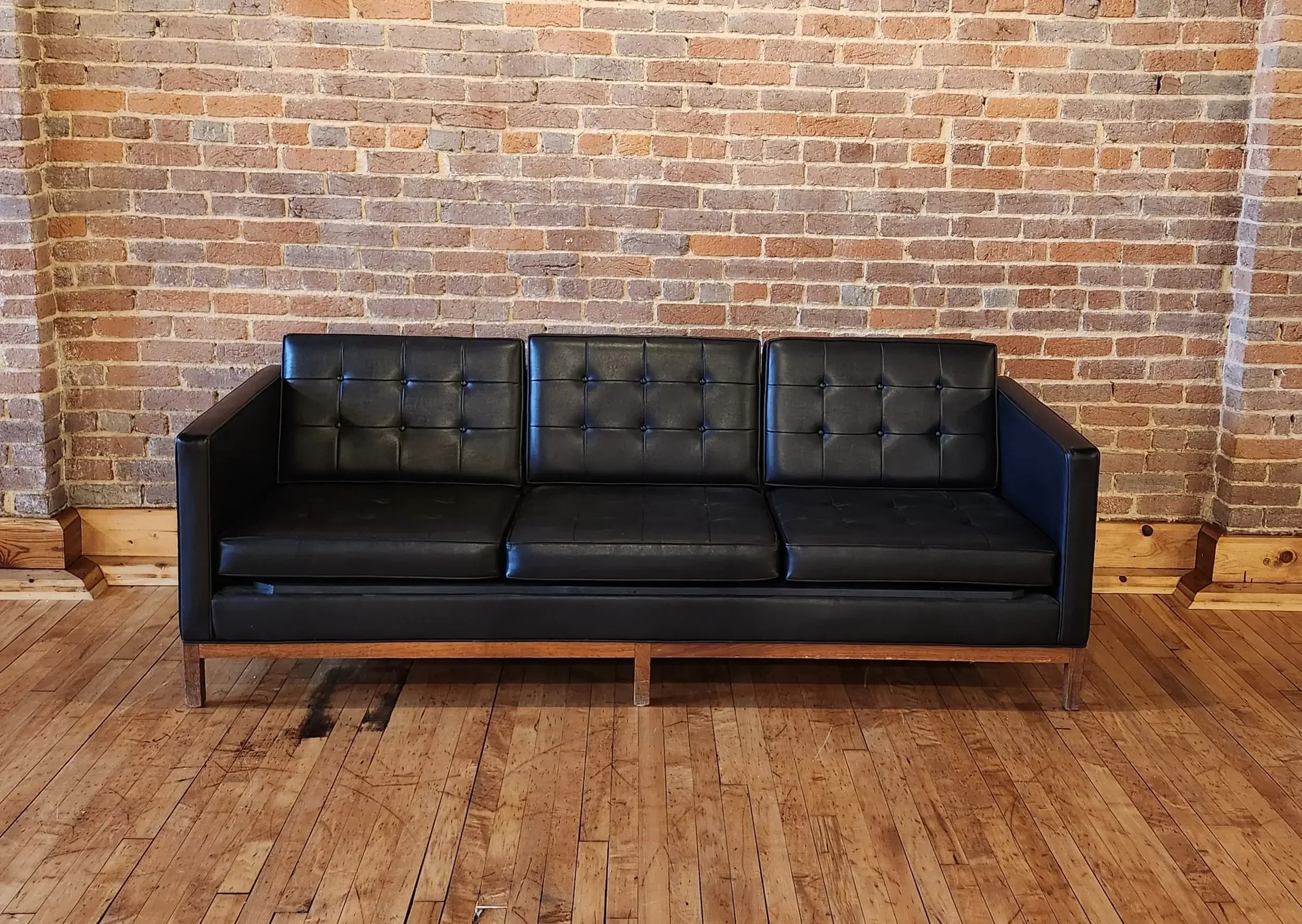 A black leather couch with a wooden frame and three padded seats, positioned against a brick wall in a room with wooden flooring.