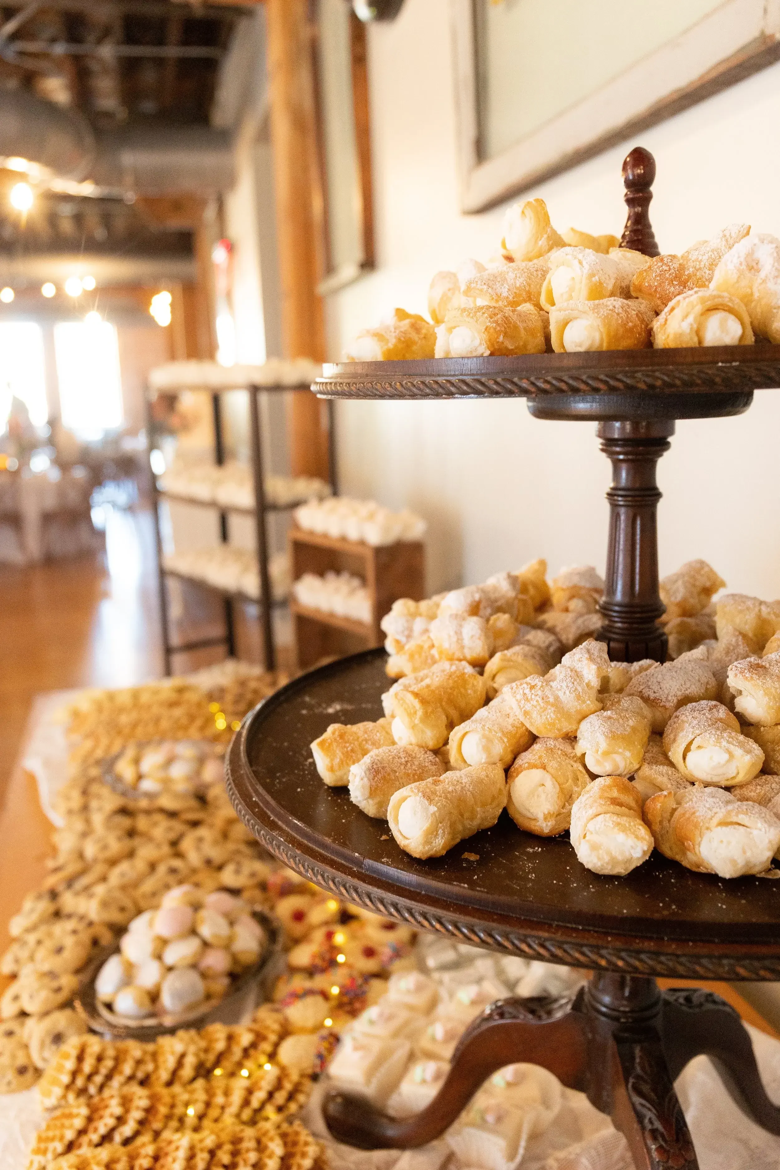 A dessert table with a two-tiered stand filled with cannoli, powdered sugar on top, in a cozy, rustic venue with blurred string lights and additional baked goods in the background.