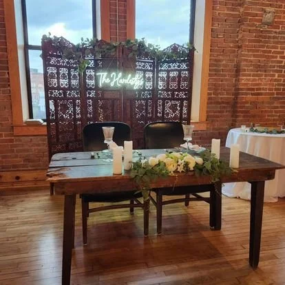 Rustic wooden table decorated with white flowers and candles, set inside a brick-walled room with large windows.