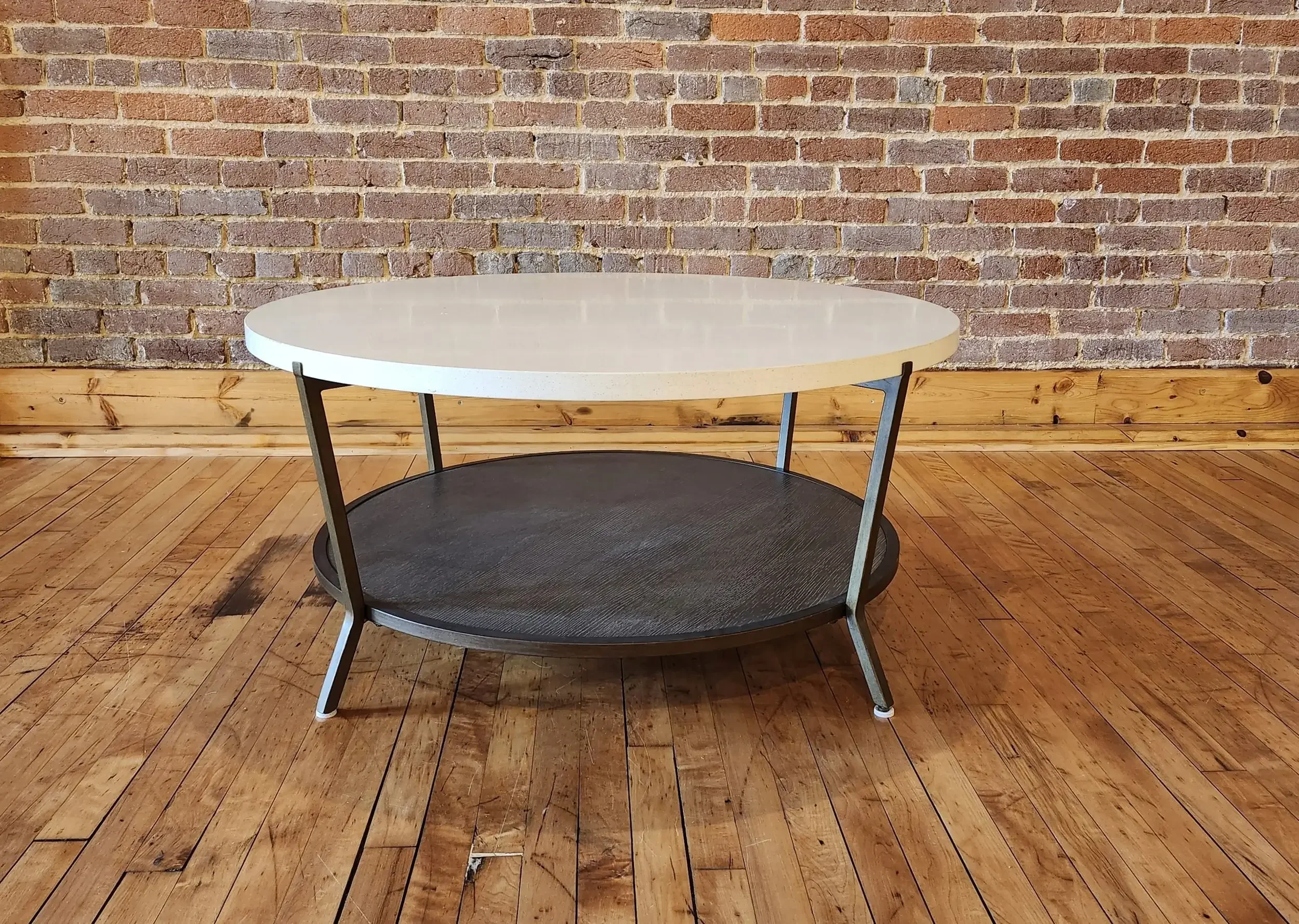 A round coffee table with a white top, black middle shelf, and angled metal legs, placed on wooden floorboards against an exposed brick wall.