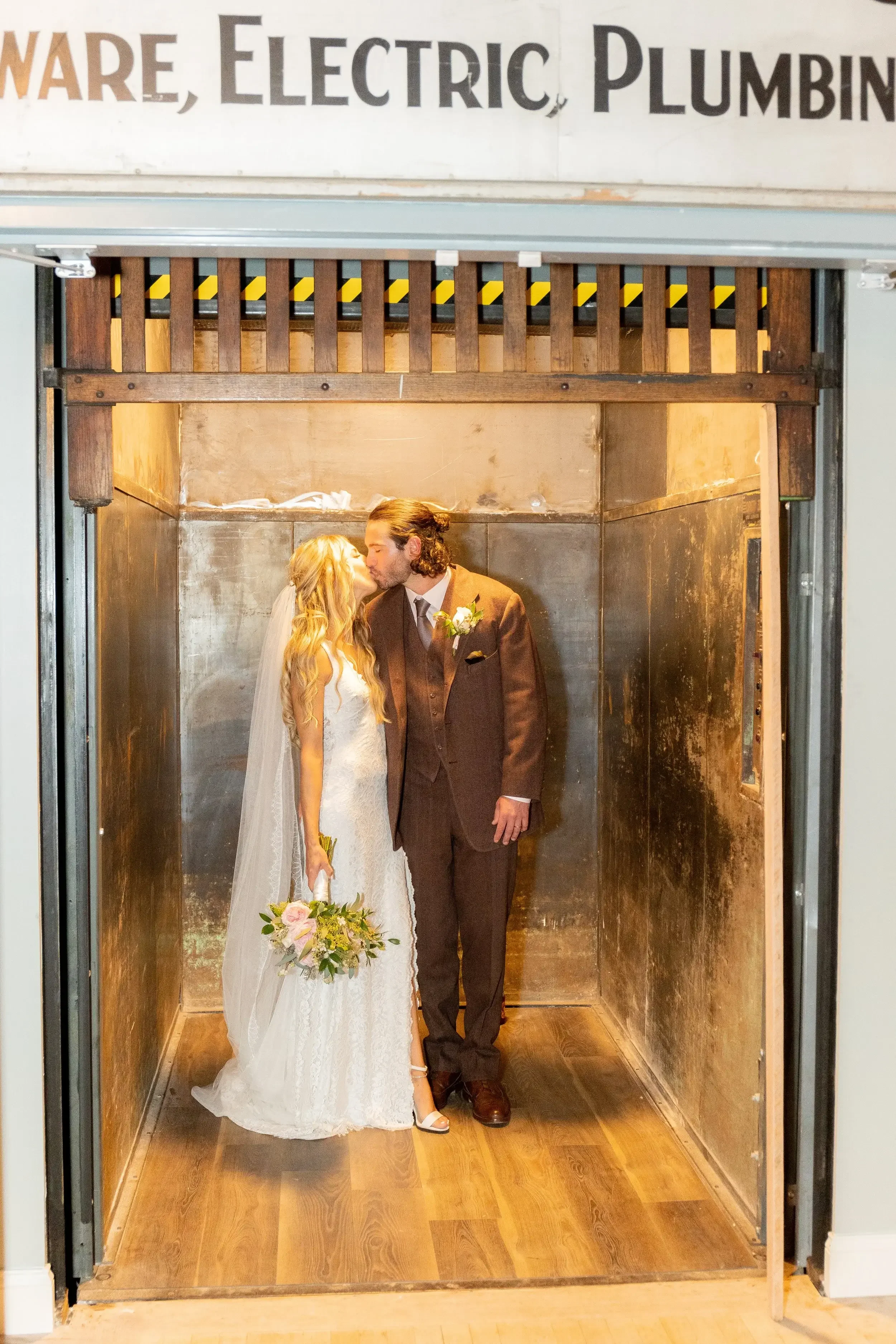 A bride and groom sharing a kiss inside a vintage elevator with metal walls and wooden flooring.