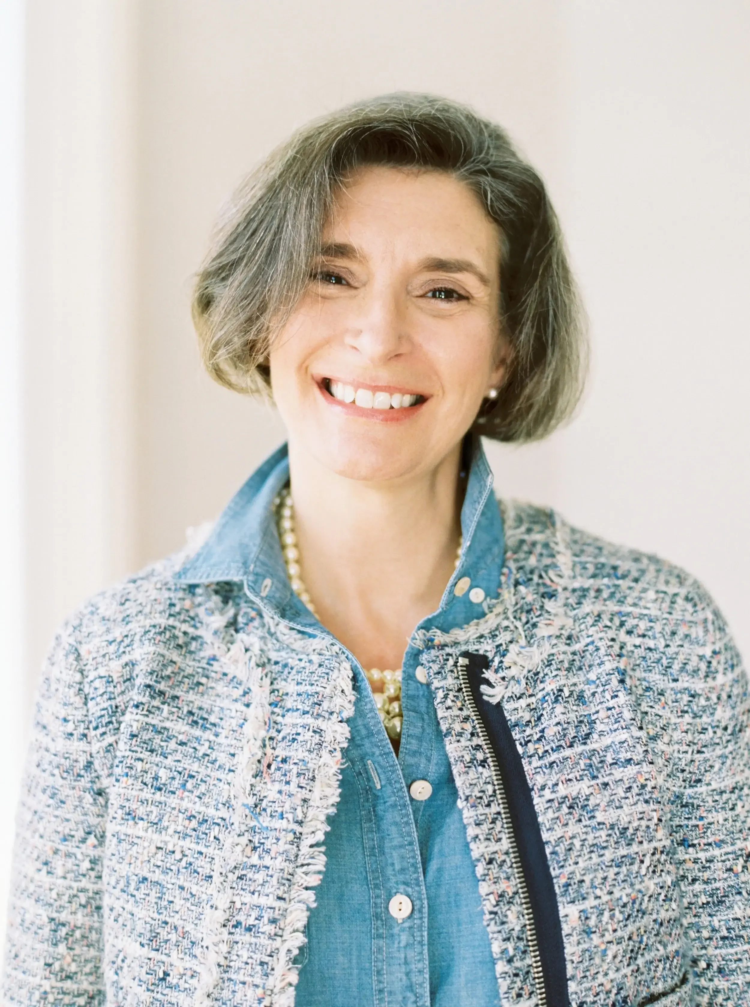 A woman with short, gray hair smiling, wearing a pearl necklace, stud earrings, a blue denim shirt, and a multicolored tweed jacket.