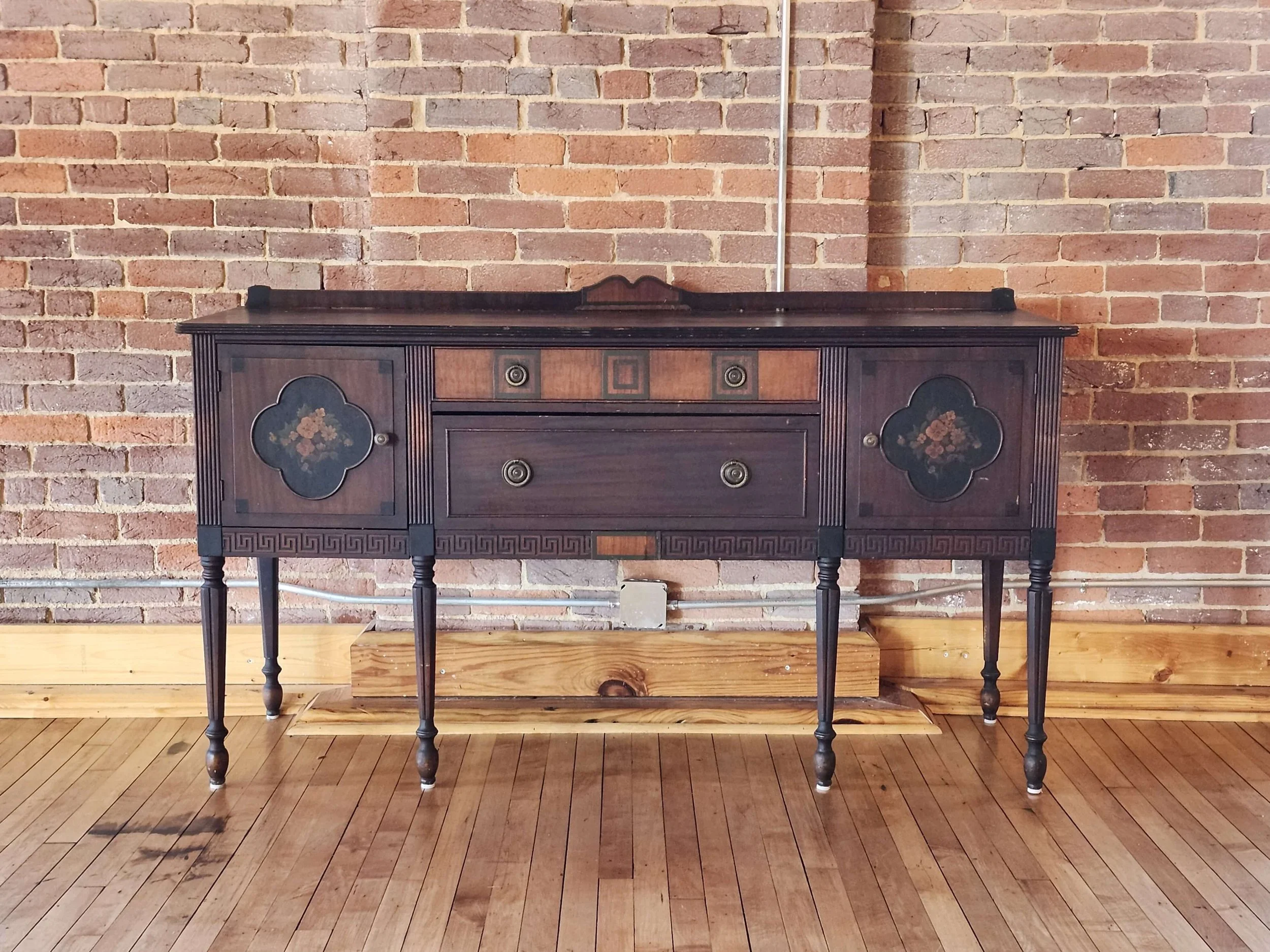 Vintage black wooden sideboard with floral details on the doors, set against a brick wall on a hardwood floor.