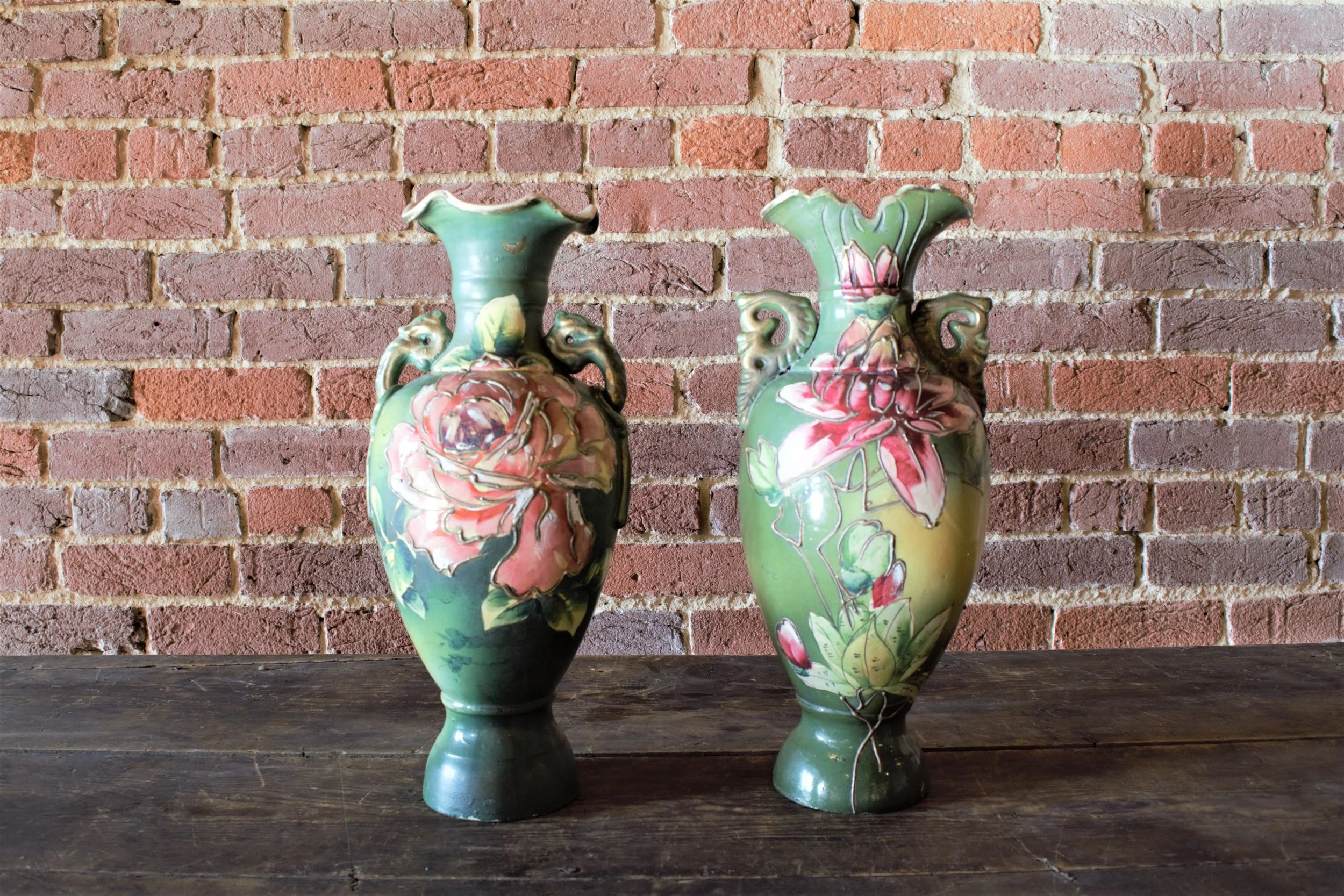 Two decorative ceramic vases with floral patterns sitting on a dark wooden surface against a brick wall background.
